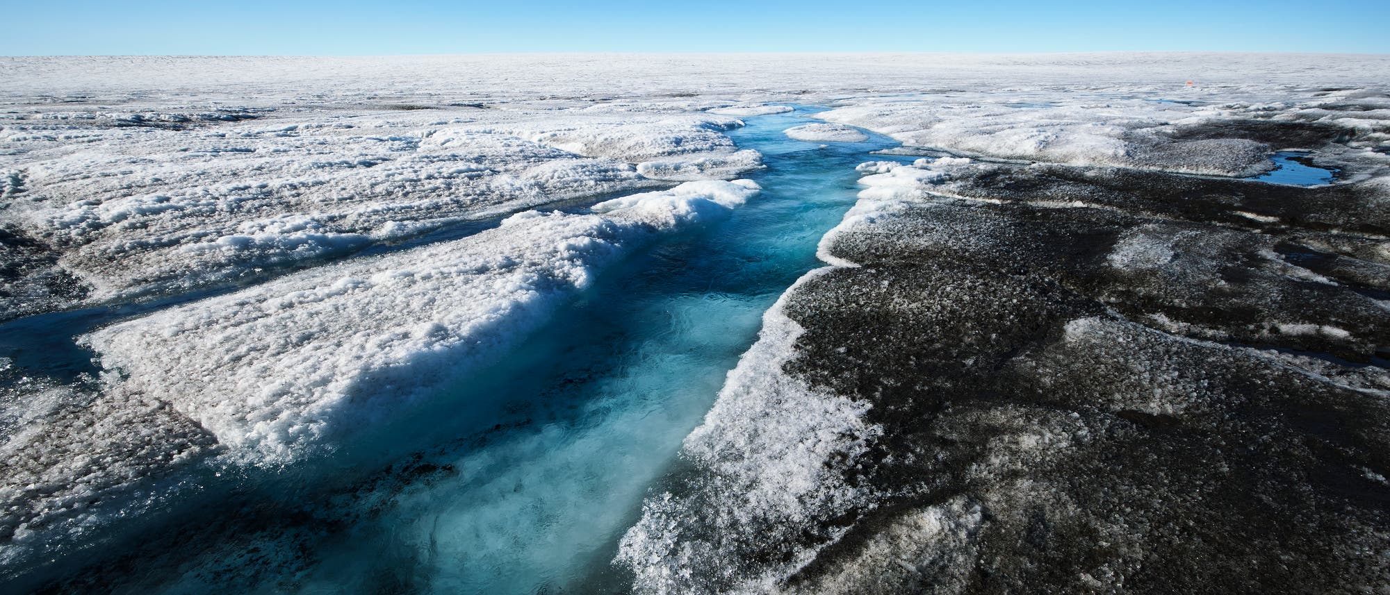 Eine weite, eisbedeckte Landschaft mit einem schmalen, blauen Schmelzwasserfluss, der sich durch das Eis schlängelt. Der Himmel ist klar und blau, was die kühle, arktische Umgebung unterstreicht. Die Oberfläche des Eises zeigt dunkle und helle Bereiche, die die Textur und Struktur des gefrorenen Terrains hervorheben. Eine weite, eisbedeckte Landschaft mit einem schmalen, blauen Schmelzwasserfluss, der sich durch das Eis schlängelt. Der Himmel ist klar und blau, was die kühle, arktische Umgebung unterstreicht. Die Oberfläche des Eises zeigt dunkle und helle Bereiche, die die Textur und Struktur des gefrorenen Terrains hervorheben.