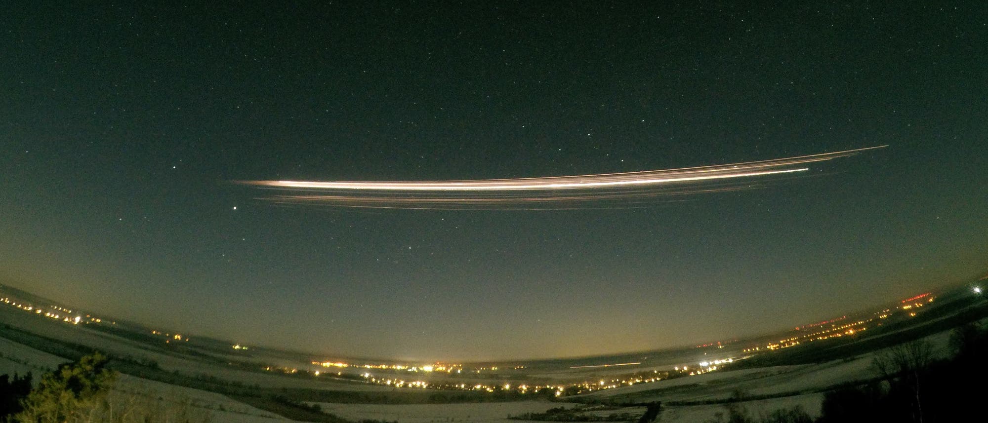 Eine n&auml;chtliche Landschaft mit einem klaren Sternenhimmel. Ein heller Lichtstreifen zieht sich horizontal &uuml;ber den Himmel, vermutlich ein Meteor oder ein Satellit. Unten sind Lichter einer Stadt oder Siedlung zu sehen, die die dunkle Landschaft erhellen. Der Himmel ist mit zahlreichen Sternen &uuml;bers&auml;t.