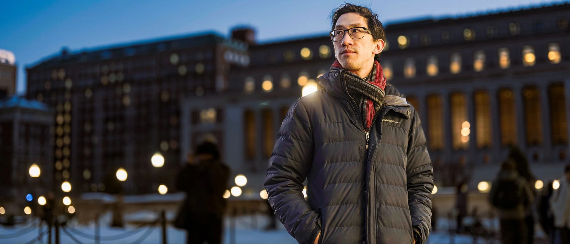 Eine Person steht in einem winterlichen Stadtpark bei D&auml;mmerung. Sie tr&auml;gt eine dicke Jacke und einen Schal. Im Hintergrund sind beleuchtete Geb&auml;ude und Stra&szlig;enlaternen zu sehen, die eine warme Atmosph&auml;re schaffen. Der Boden ist teilweise mit Schnee bedeckt, und einige Menschen gehen im Hintergrund entlang.
