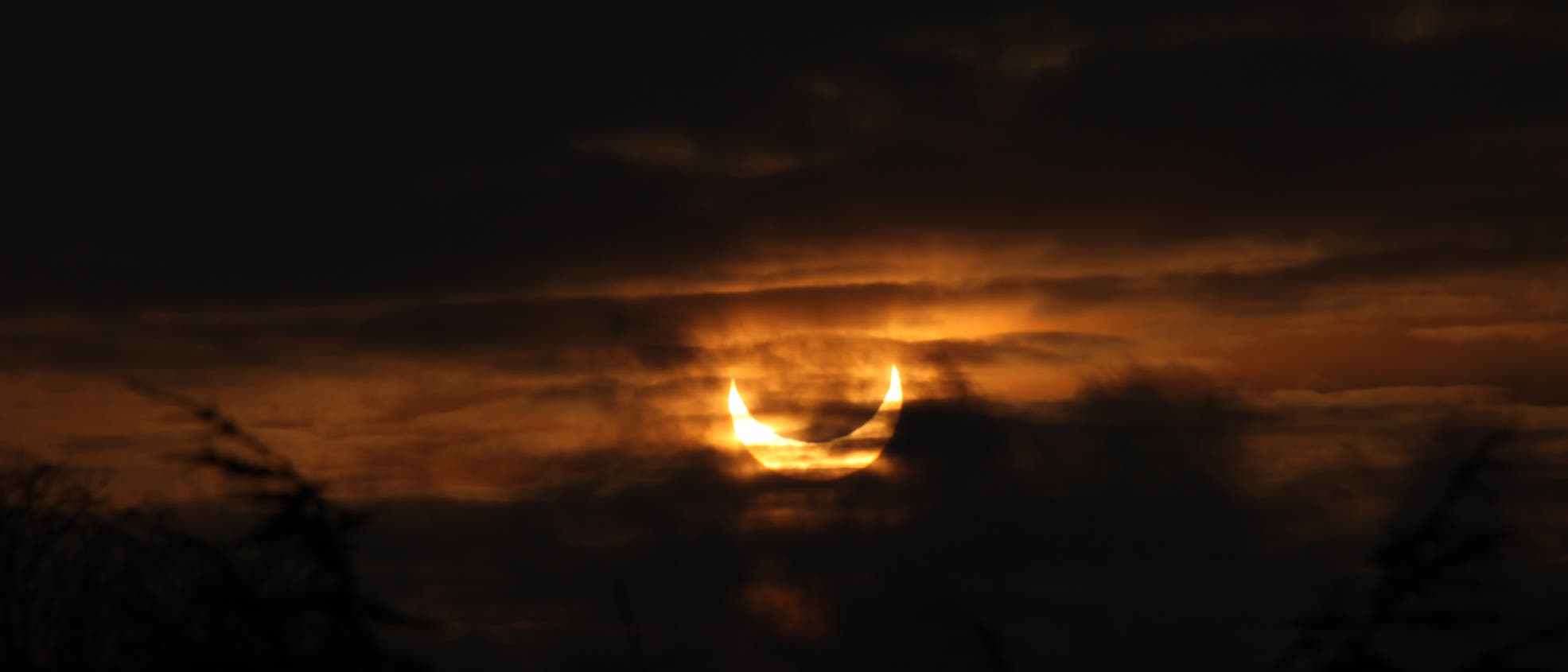 Eine partielle Sonnenfinsternis am Himmel, bei der die Sonne teilweise vom Mond verdeckt wird. Der Himmel ist dunkel mit einigen Wolken, die die leuchtende Sichel der Sonne umgeben. Die Szene vermittelt eine mystische Atmosphäre. Im Vordergrund sind Silhouetten von Baumspitzen zu sehen. Eine partielle Sonnenfinsternis am Himmel, bei der die Sonne teilweise vom Mond verdeckt wird. Der Himmel ist dunkel mit einigen Wolken, die die leuchtende Sichel der Sonne umgeben. Die Szene vermittelt eine mystische Atmosphäre. Im Vordergrund sind Silhouetten von Baumspitzen zu sehen.