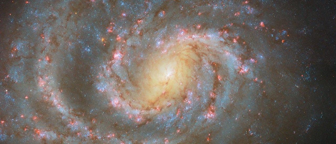 Eine Spiralgalaxie mit einem hellen, leuchtenden Zentrum und ausgedehnten, spiralförmigen Armen, die mit blauen und roten Sternen übersät sind. Die Galaxie ist von einem dunklen Weltraumhintergrund umgeben, in dem vereinzelt weitere Sterne zu sehen sind. Die Struktur der Galaxie zeigt deutlich die spiralförmige Anordnung der Sterne und interstellaren Materie.