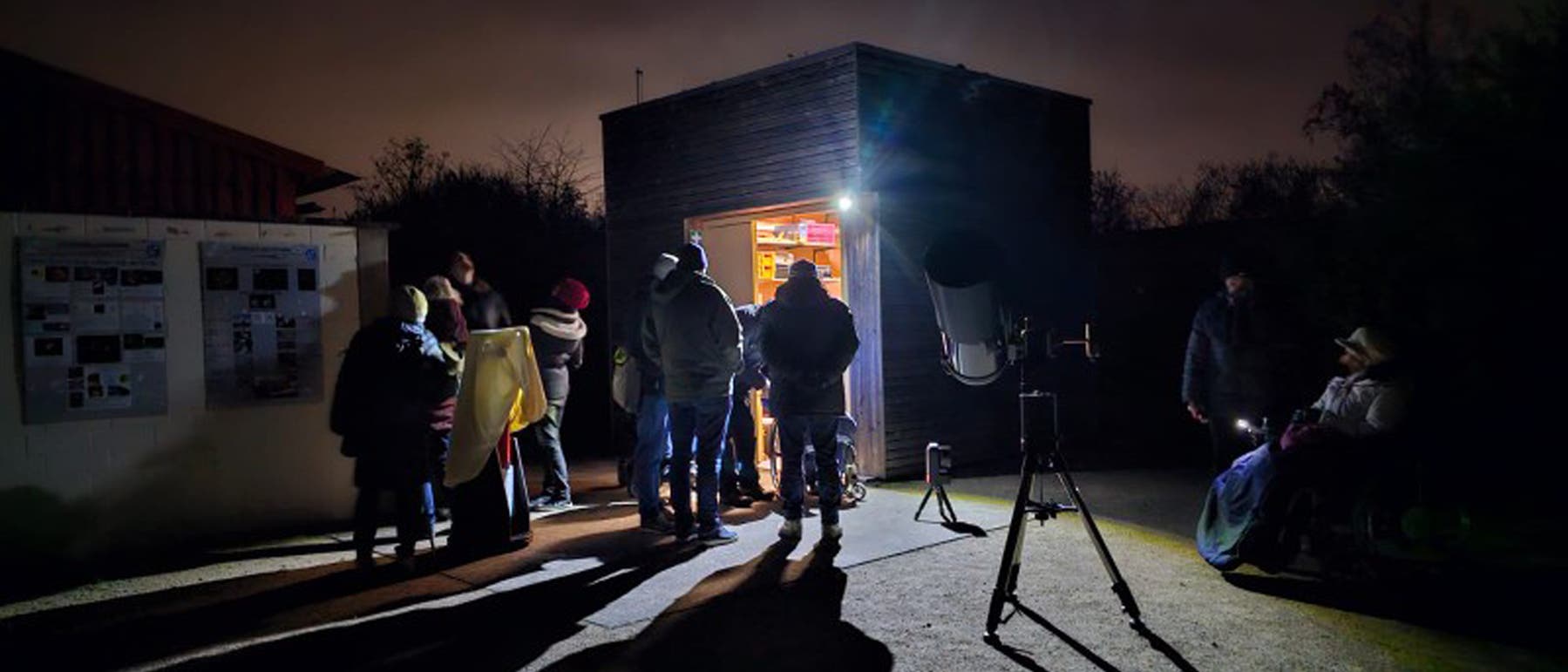 Varias personas permanecen en la oscuridad en un pequeño cuadrado frente a un cobertizo de equipo, desde el cual ilumina la luz. Se establecen varios telescopios que son examinados por los presentes. Varias personas permanecen en la oscuridad en un pequeño cuadrado frente a un cobertizo de equipo, desde el cual ilumina la luz. Se establecen varios telescopios que son examinados por los presentes.