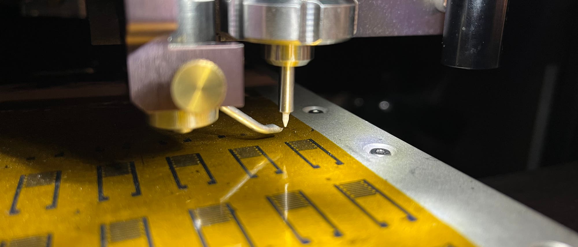 Nahaufnahme einer 3-D-Druckerdüse, die auf einer gelben Leiterplatte arbeitet. Auf der Leiterplatte sind mehrere schwarze rechteckige Muster zu sehen. Die Szene ist gut beleuchtet, was die Details der Maschine und der Leiterplatte hervorhebt. Nahaufnahme einer 3-D-Druckerdüse, die auf einer gelben Leiterplatte arbeitet. Auf der Leiterplatte sind mehrere schwarze rechteckige Muster zu sehen. Die Szene ist gut beleuchtet, was die Details der Maschine und der Leiterplatte hervorhebt.