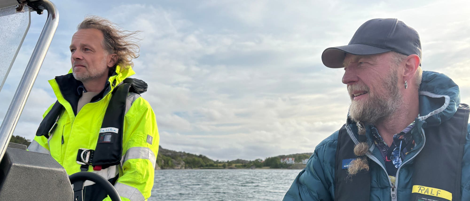 Zwei Personen stehen nebeneinander auf einem Boot. Beide tragen wetterfeste Jacken und Schwimmwesten. Im Vordergrund ist ein Teil eines Bootes mit der Aufschrift "Radar Reflector" sichtbar. Der Himmel ist bewölkt. Die Personen lächeln und scheinen sich in einer maritimen Umgebung zu befinden.