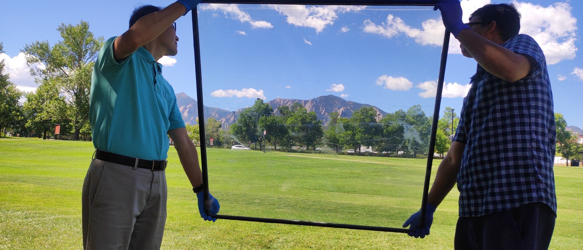 Zwei Personen stehen im Freien auf einer Wiese und halten einen gro&szlig;en, rechteckigen Glasrahmen. Der Himmel ist klar und blau mit einigen Wolken, und im Hintergrund sind Berge zu sehen. Die Personen tragen Handschuhe und scheinen den Rahmen zu inspizieren oder zu pr&auml;sentieren. Die Szene vermittelt einen Eindruck von einem sonnigen Tag in einer landschaftlich reizvollen Umgebung.
