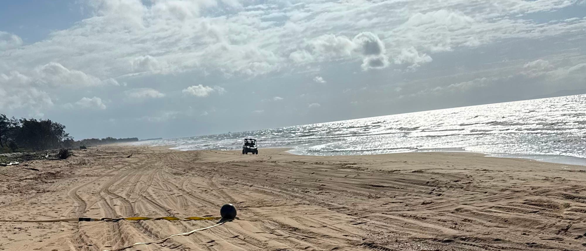 Paisaje costero con un vehículo todoterreno circulando por una costa arenosa. El cielo está nublado y el mar brilla a la luz del sol. En primer plano hay una cuerda con una pelota en la arena. Paisaje costero con un vehículo todoterreno circulando por una costa arenosa. El cielo está nublado y el mar brilla a la luz del sol. En primer plano hay una cuerda con una pelota en la arena.