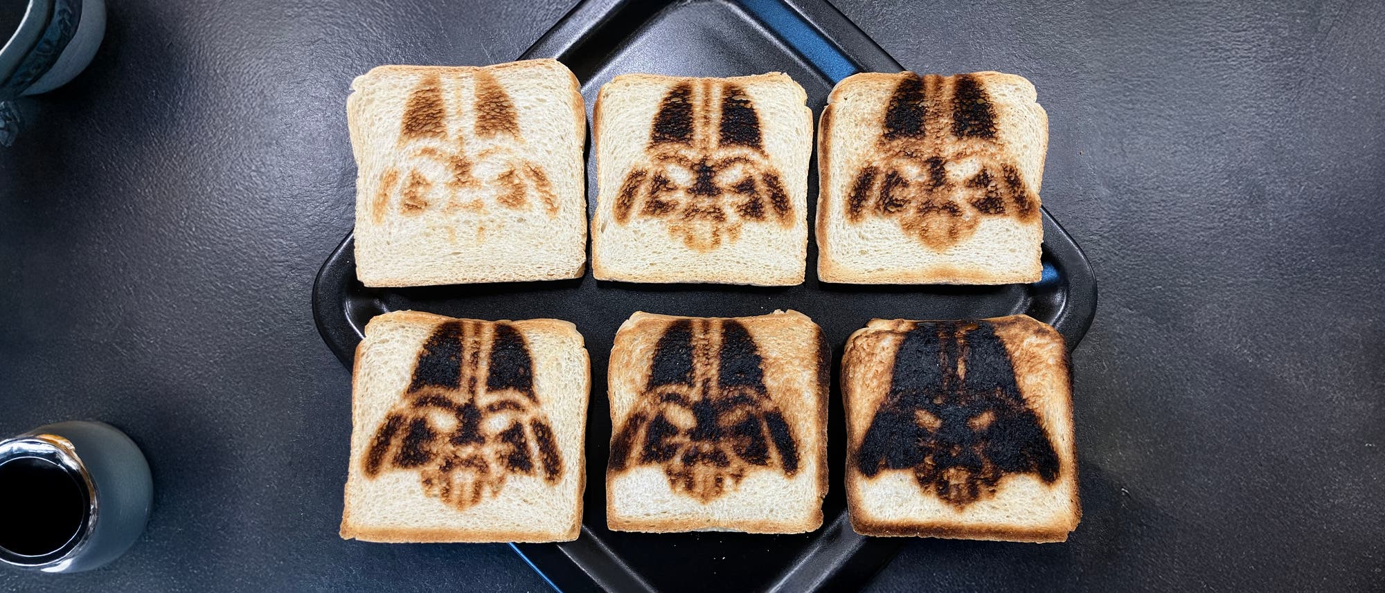 Sechs Scheiben Toastbrot auf einem schwarzen Tablett, jede mit einem dunklen, gebräunten Muster, das einem ikonischen Helm ähnelt. Links unten ist eine kleine, schwarze Tasse zu sehen. Der Hintergrund ist eine dunkle Oberfläche. Sechs Scheiben Toastbrot auf einem schwarzen Tablett, jede mit einem dunklen, gebräunten Muster, das einem ikonischen Helm ähnelt. Links unten ist eine kleine, schwarze Tasse zu sehen. Der Hintergrund ist eine dunkle Oberfläche.