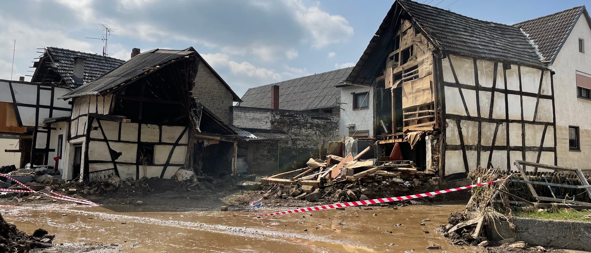 Ein zerst&ouml;rtes Fachwerkhaus in einer &uuml;berfluteten Stra&szlig;e. Tr&uuml;mmer und Schlamm bedecken den Boden, w&auml;hrend Absperrband den Bereich sichert. Der Himmel ist bew&ouml;lkt, und die Szene vermittelt den Eindruck einer k&uuml;rzlich erfolgten Naturkatastrophe.