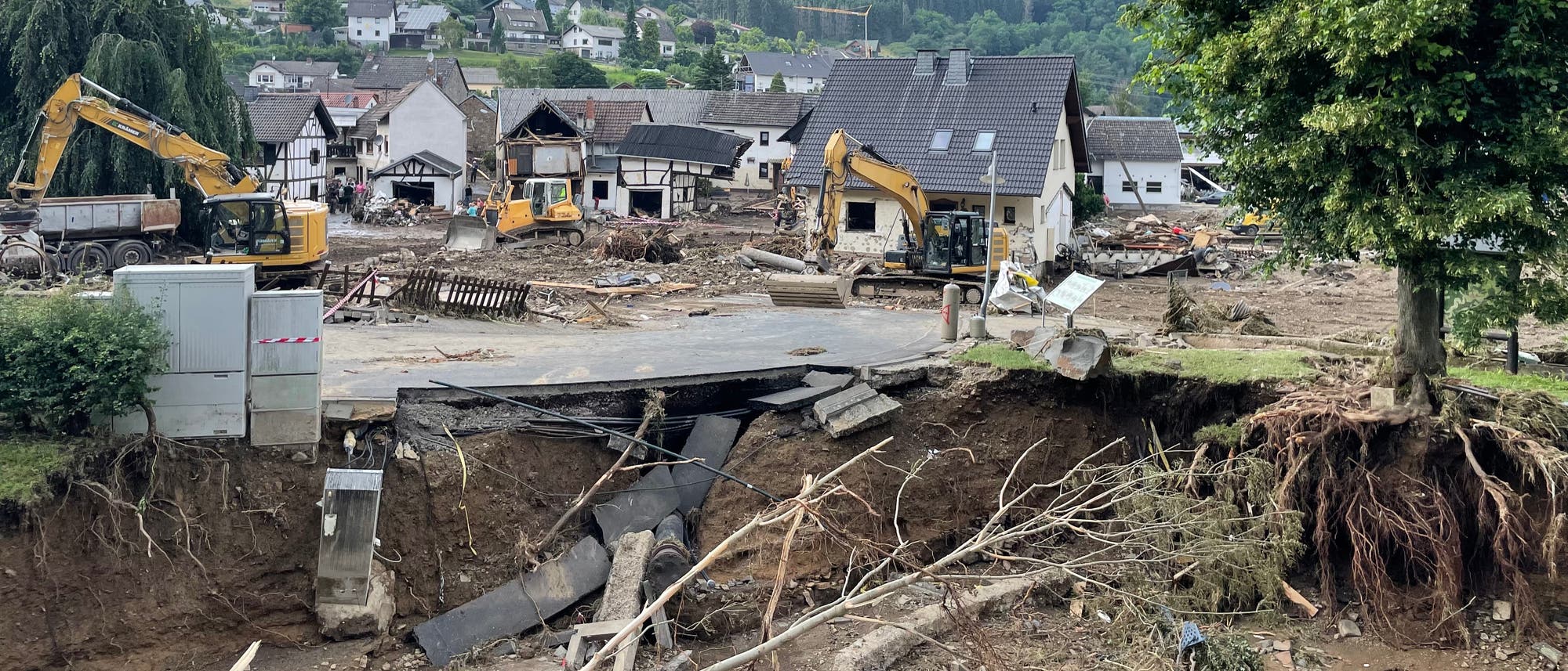 Eine Szene von Hochwassersch&auml;den in einem Dorf. Mehrere Bagger sind im Einsatz, um Tr&uuml;mmer zu beseitigen. Ein Haus mit besch&auml;digtem Dach steht im Hintergrund, umgeben von zerst&ouml;rten Geb&auml;uden und umgest&uuml;rzten B&auml;umen. Der Boden ist erodiert, und freiliegende Wurzeln und Rohre sind sichtbar. Im Vordergrund ist eine abgebrochene Stra&szlig;e zu sehen. Die Umgebung zeigt eine h&uuml;gelige Landschaft mit weiteren H&auml;usern im Hintergrund.