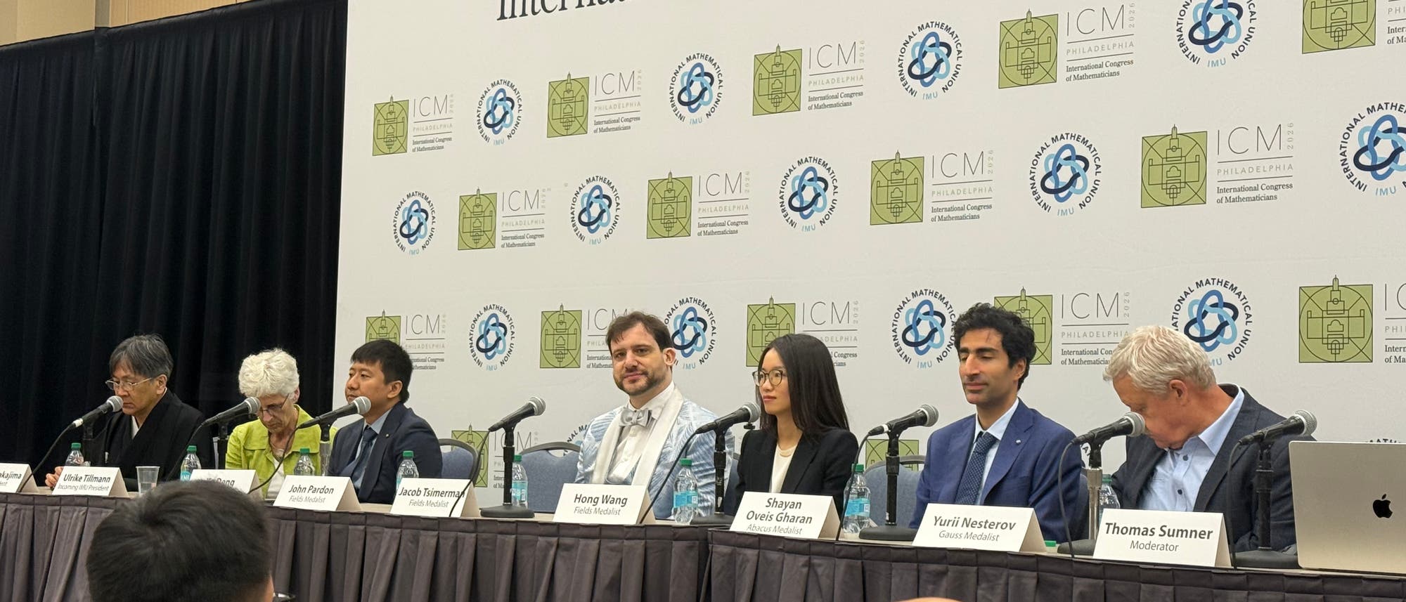 Eine Gruppe von Personen sitzt an einem Tisch während einer Podiumsdiskussion auf einer Konferenz. Hinter ihnen ist eine Wand mit Logos des International Congress of Mathematicians (ICM) und der International Mathematical Union (IMU) zu sehen. Vor jedem Teilnehmer steht ein Mikrofon, und Namensschilder identifizieren sie als Moderatoren und Preisträger. Die Atmosphäre ist formell und professionell. Eine Gruppe von Personen sitzt an einem Tisch während einer Podiumsdiskussion auf einer Konferenz. Hinter ihnen ist eine Wand mit Logos des International Congress of Mathematicians (ICM) und der International Mathematical Union (IMU) zu sehen. Vor jedem Teilnehmer steht ein Mikrofon, und Namensschilder identifizieren sie als Moderatoren und Preisträger. Die Atmosphäre ist formell und professionell.