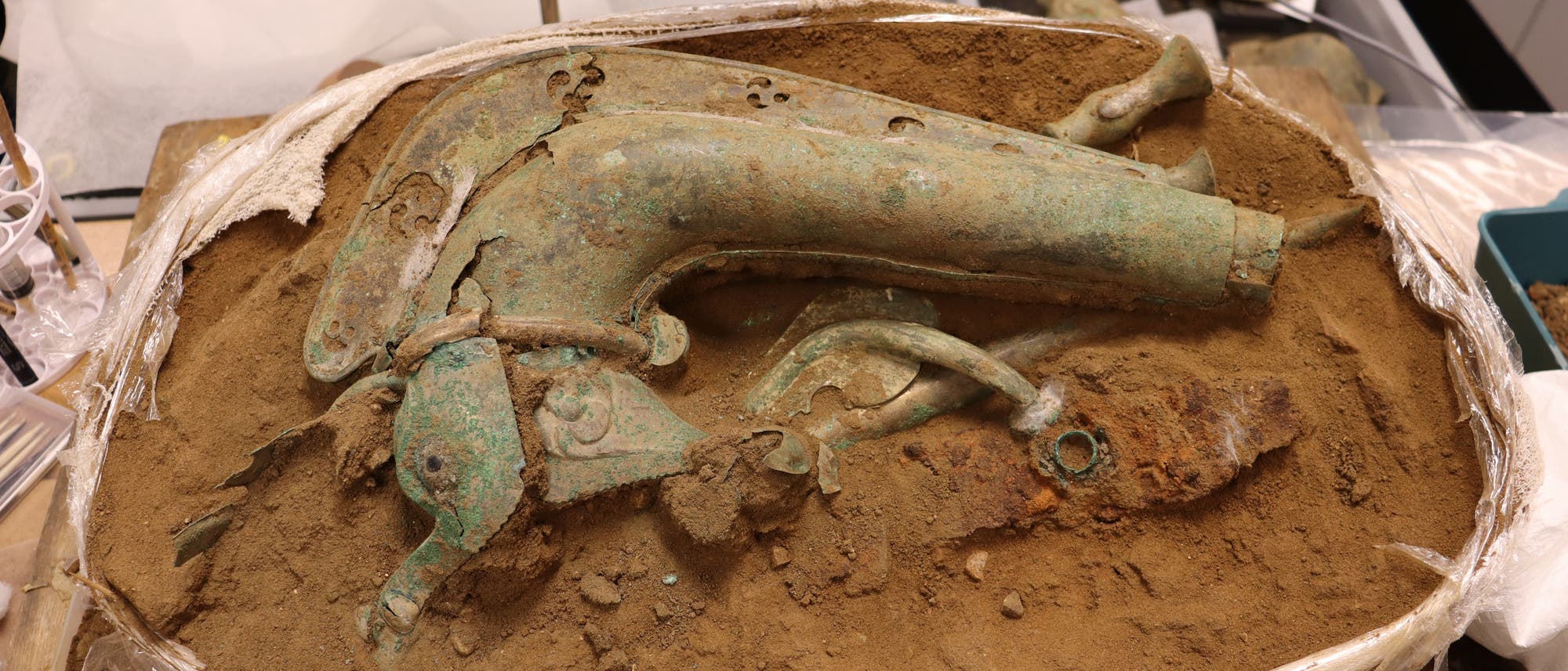 Ein antikes, teilweise freigelegtes Artefakt aus Metall, das in einem rechteckigen Beh&auml;lter mit Erde liegt. Das Objekt hat eine geschwungene Form und zeigt Anzeichen von Korrosion. Im Hintergrund sind weitere arch&auml;ologische Funde und Werkzeuge zu sehen. Das Bild vermittelt den Eindruck einer arch&auml;ologischen Ausgrabung oder Restaurierung.