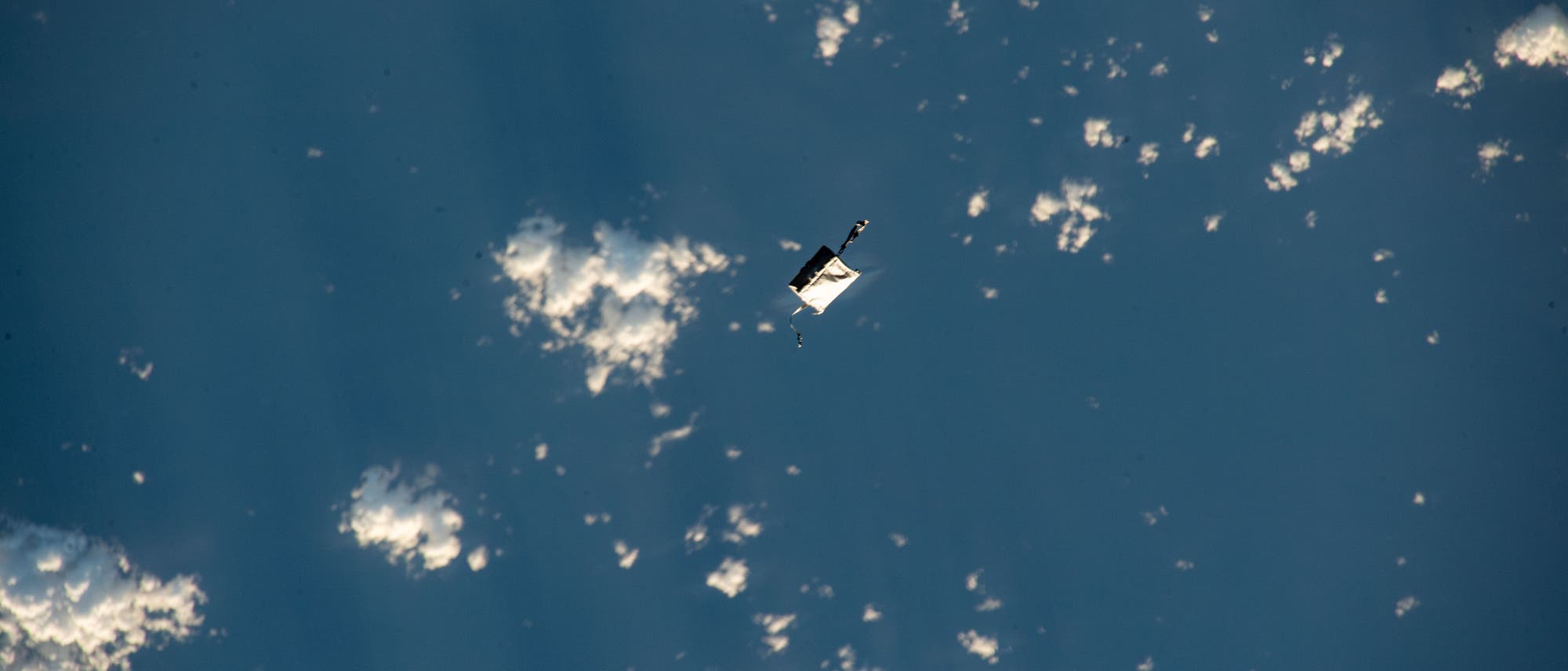 Eine Werkzeugtasche in Weiß fliegt durch das All und überquert einen blauen Ozean mit Wolken am Himmel.