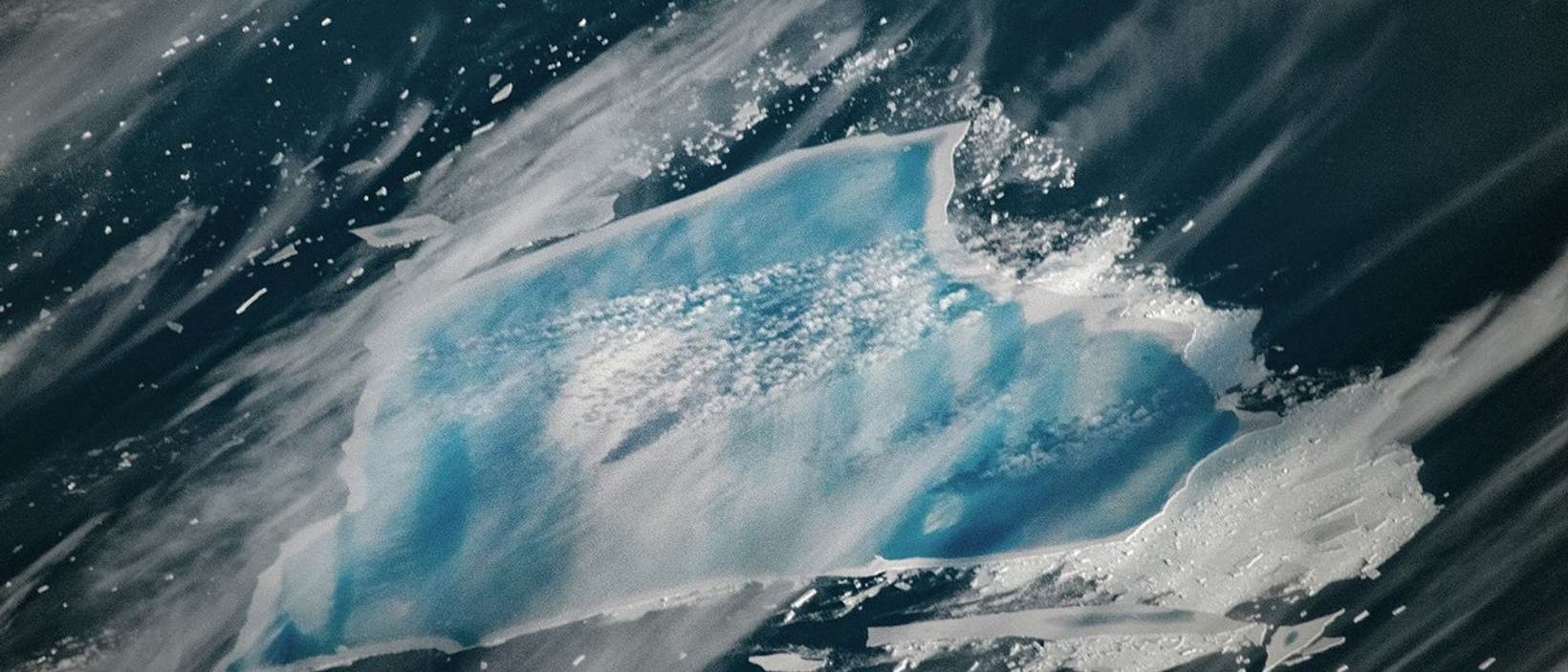 Luftaufnahme eines gro&szlig;en, unregelm&auml;&szlig;ig geformten Eisbergs, der im dunklen Wasser treibt. Der Eisberg ist von schmelzendem Eis und kleineren Eisschollen umgeben. Die Oberfl&auml;che des Eisbergs zeigt eine Mischung aus hellem Blau und Wei&szlig;, was auf die unterschiedlichen Eisschichten hinweist. Die Szene vermittelt ein Gef&uuml;hl von K&auml;lte und Isolation.