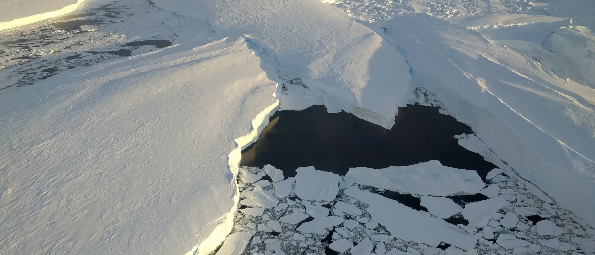 Luftaufnahme einer riesigen, schneebedeckten Eisfläche in der Antarktis, die sich bis zum Horizont erstreckt. In der Mitte des Bildes ist ein großer Riss zu sehen, der mit dunklem Wasser gefüllt ist und von zerbrochenen Eisschollen umgeben ist. Die Szene wird von der tiefstehenden Sonne beleuchtet, die lange Schatten auf das Eis wirft. Luftaufnahme einer riesigen, schneebedeckten Eisfläche in der Antarktis, die sich bis zum Horizont erstreckt. In der Mitte des Bildes ist ein großer Riss zu sehen, der mit dunklem Wasser gefüllt ist und von zerbrochenen Eisschollen umgeben ist. Die Szene wird von der tiefstehenden Sonne beleuchtet, die lange Schatten auf das Eis wirft.