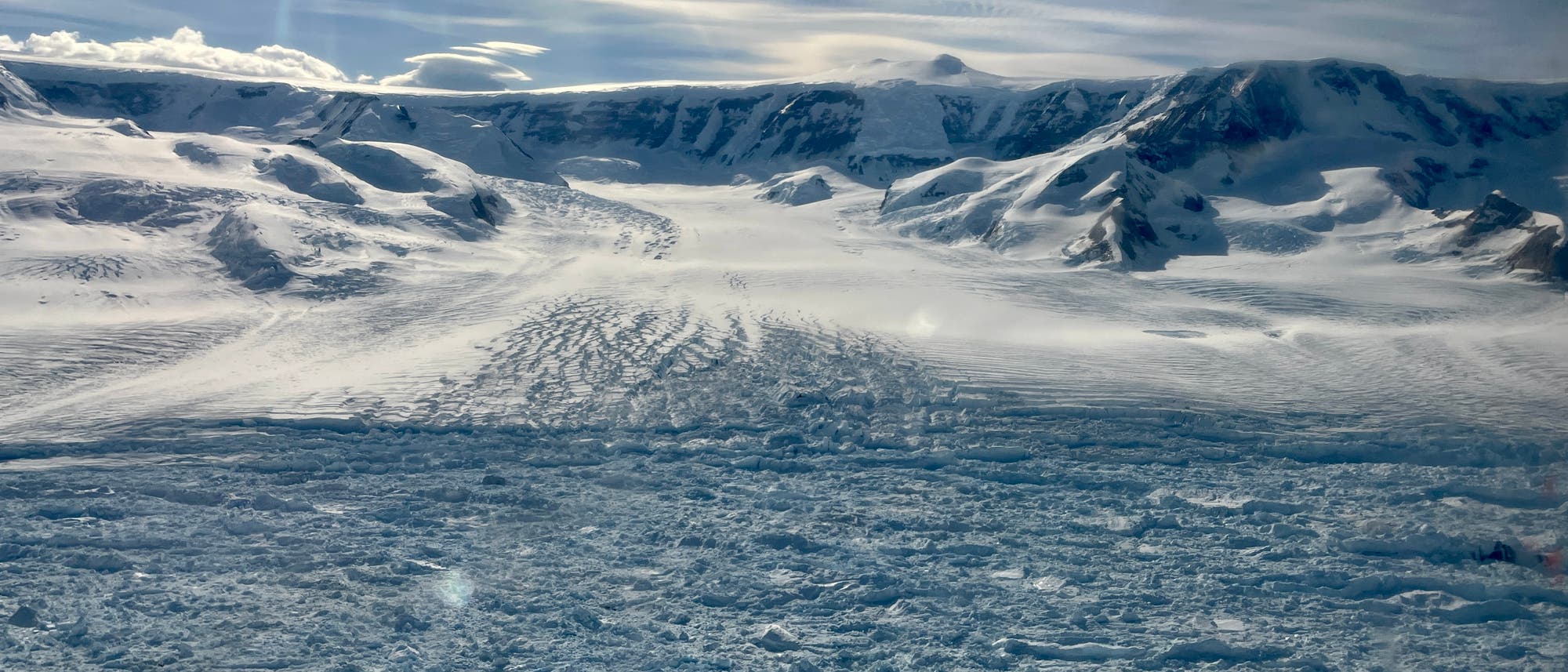 Eine weite, verschneite Landschaft mit einem Gletscher, der sich durch ein Tal zwischen schneebedeckten Bergen erstreckt. Der Himmel ist bewölkt, und Sonnenstrahlen brechen durch die Wolken, was der Szene eine dramatische Beleuchtung verleiht. Die Oberfläche des Gletschers zeigt Risse und Unebenheiten, die die Bewegung des Eises andeuten. Eine weite, verschneite Landschaft mit einem Gletscher, der sich durch ein Tal zwischen schneebedeckten Bergen erstreckt. Der Himmel ist bewölkt, und Sonnenstrahlen brechen durch die Wolken, was der Szene eine dramatische Beleuchtung verleiht. Die Oberfläche des Gletschers zeigt Risse und Unebenheiten, die die Bewegung des Eises andeuten.