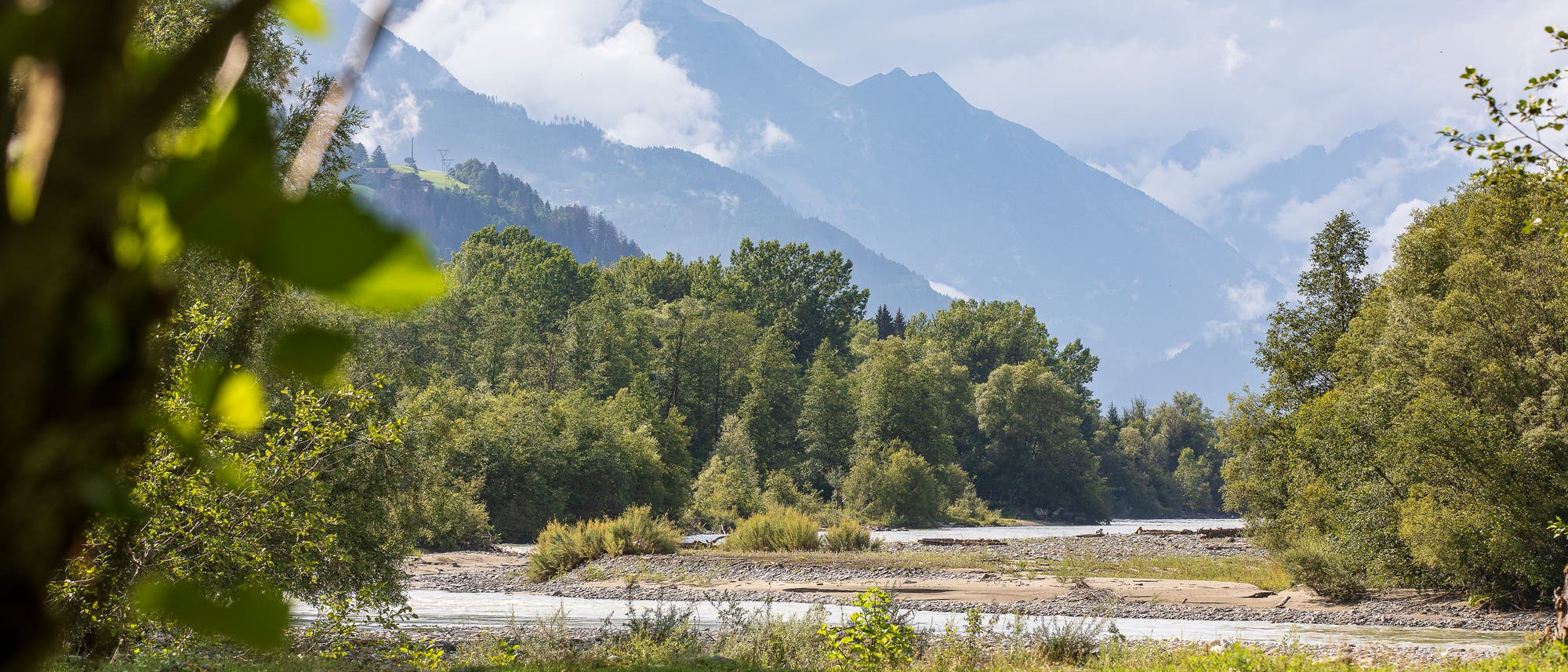 Eine malerische Landschaft mit einem Fluss im Vordergrund, umgeben von &uuml;ppigem Gr&uuml;n und B&auml;umen. Im Hintergrund erheben sich majest&auml;tische Berge, teils von Wolken umh&uuml;llt. Die Szene vermittelt eine ruhige und friedliche Atmosph&auml;re in der Natur.