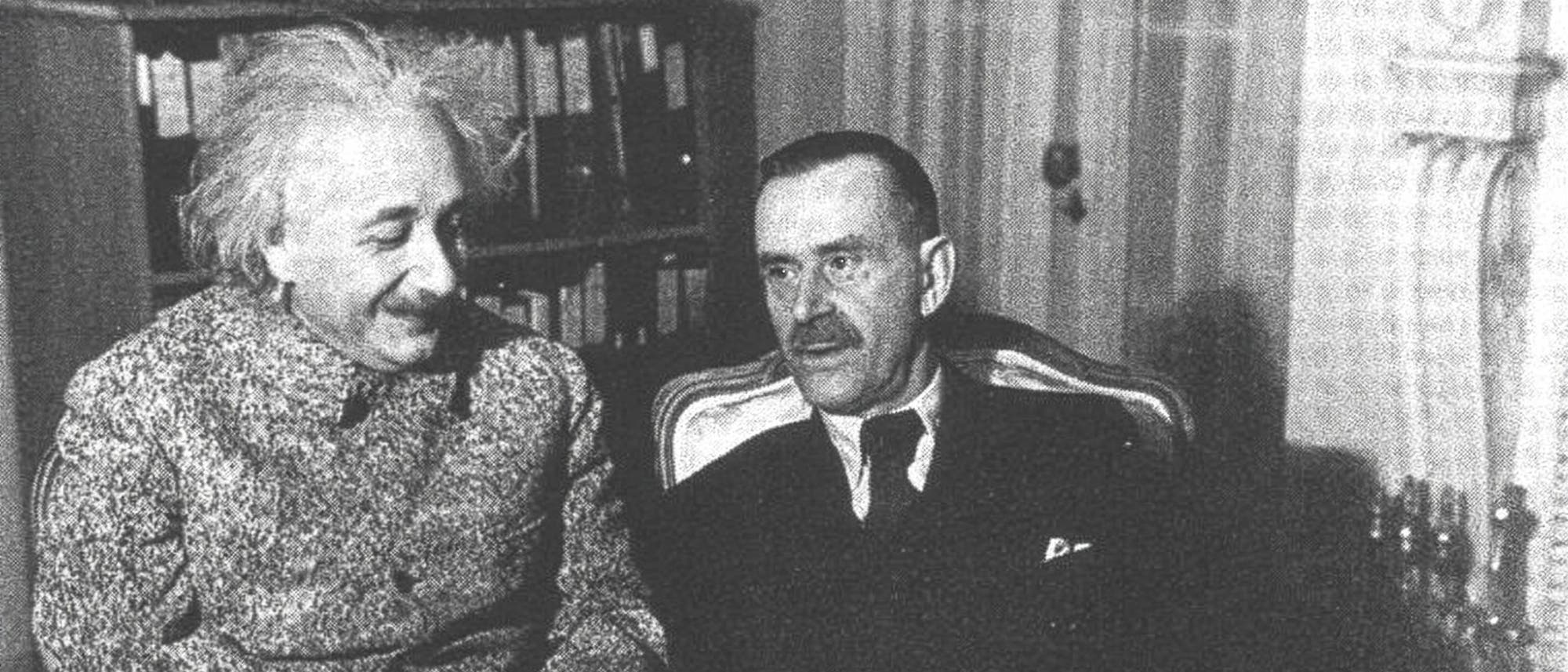 Eine &auml;ltere Schwarz-wei&szlig;-Aufnahme: Zwei M&auml;nner sitzen nebeneinander in einem Raum mit B&uuml;cherregalen im Hintergrund. Der Mann links l&auml;chelt und hat lockiges Haar, w&auml;hrend der Mann rechts einen ernsten Ausdruck hat und ein Dokument in der Hand h&auml;lt. Beide tragen Anz&uuml;ge.