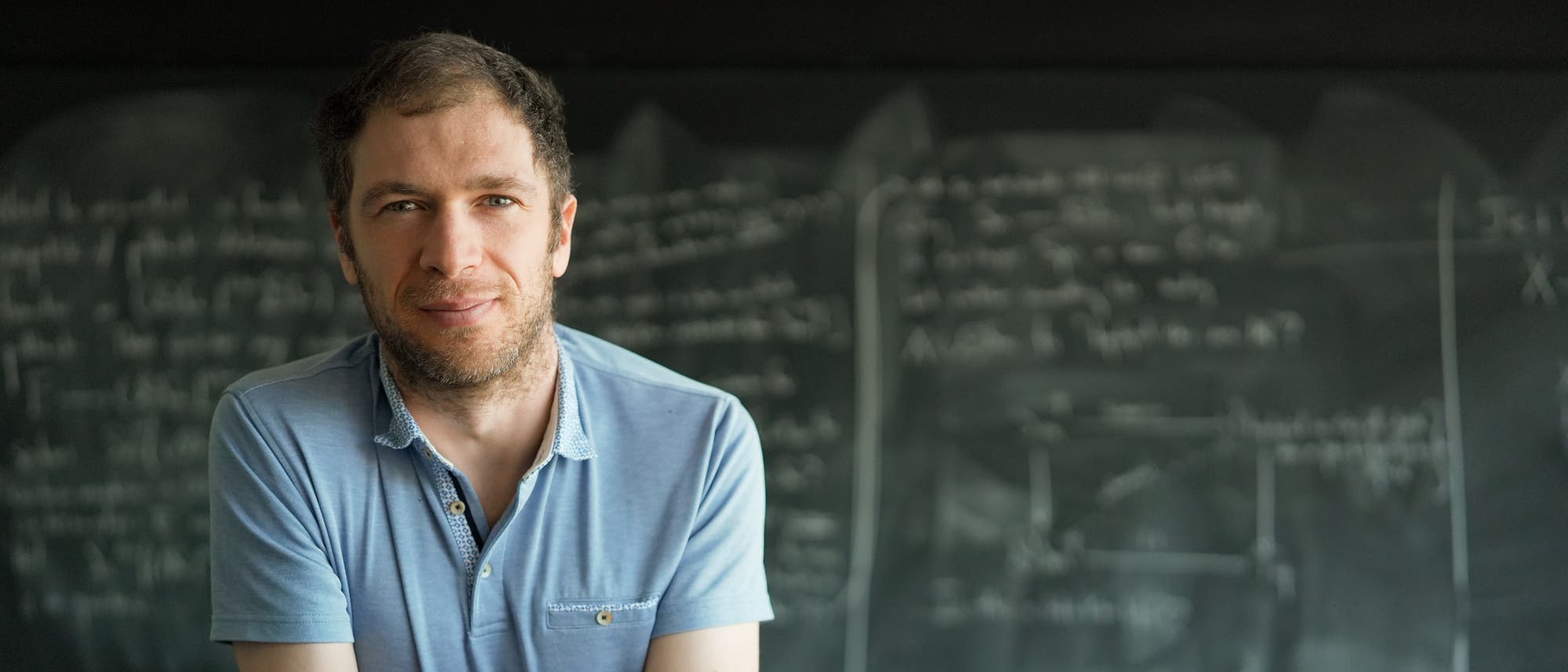 Eine Person in einem blauen Hemd steht vor einer Tafel, die mit mathematischen Formeln und Diagrammen beschrieben ist. Die Person blickt in die Kamera und wirkt nachdenklich. Die Tafel im Hintergrund ist unscharf, aber es sind komplexe Gleichungen und Skizzen erkennbar, die auf wissenschaftliche oder mathematische Arbeit hindeuten. Eine Person in einem blauen Hemd steht vor einer Tafel, die mit mathematischen Formeln und Diagrammen beschrieben ist. Die Person blickt in die Kamera und wirkt nachdenklich. Die Tafel im Hintergrund ist unscharf, aber es sind komplexe Gleichungen und Skizzen erkennbar, die auf wissenschaftliche oder mathematische Arbeit hindeuten.