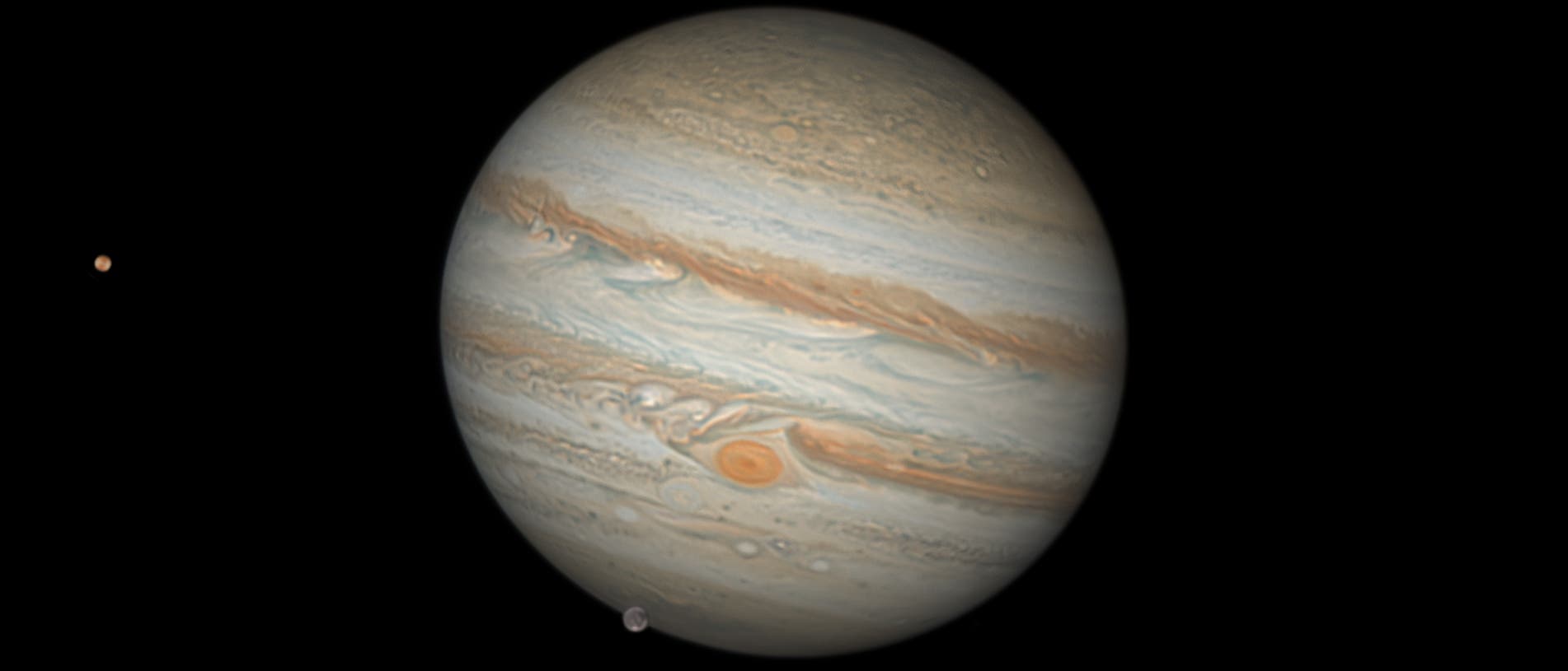 Das Bild zeigt den Planeten Jupiter mit seinen charakteristischen Wolkenb&auml;ndern und dem Gro&szlig;en Roten Fleck, einem riesigen Sturm. Zwei seiner Monde sind ebenfalls sichtbar: einer links und einer unten rechts neben dem Planeten. Der Hintergrund ist schwarz, was den Fokus auf Jupiter und seine Monde lenkt.