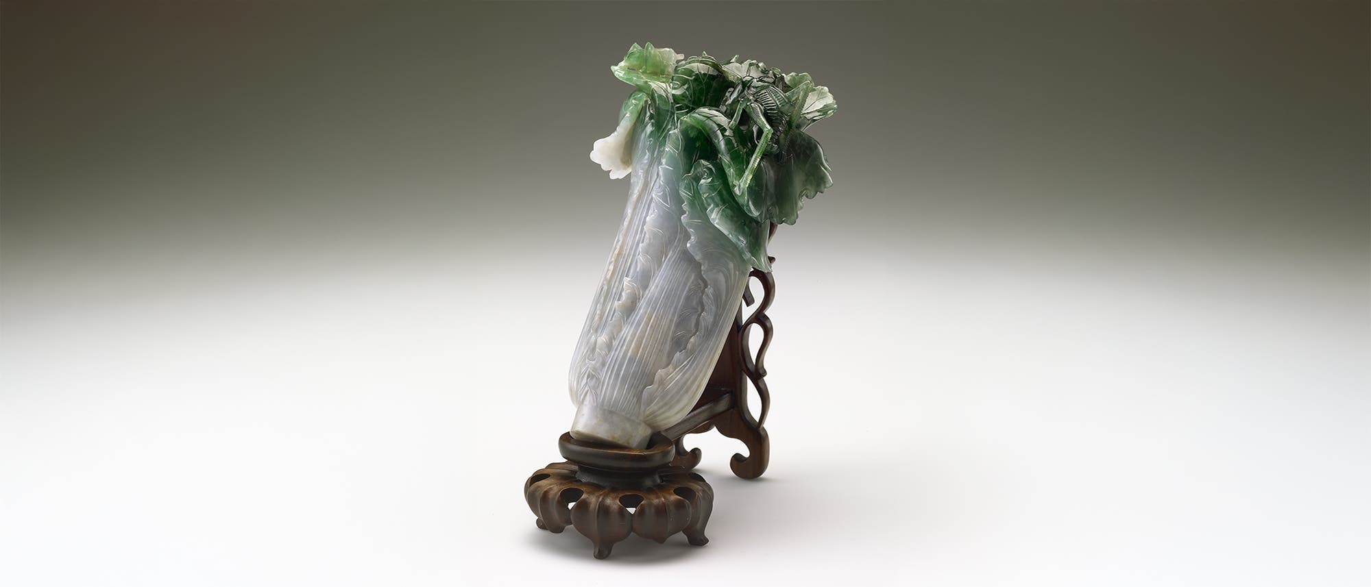 Eine kunstvolle Skulptur aus Jade, die einen chinesischen Kohl darstellt. Die Skulptur zeigt detaillierte Blätter in verschiedenen Grüntönen, die realistisch geschnitzt sind. Der Kohl ist auf einem kunstvoll gestalteten Holzständer platziert, der die Skulptur elegant präsentiert. Die Kombination aus den natürlichen Farben der Jade und der feinen Schnitzarbeit verleiht dem Kunstwerk eine lebendige und authentische Erscheinung. Eine kunstvolle Skulptur aus Jade, die einen chinesischen Kohl darstellt. Die Skulptur zeigt detaillierte Blätter in verschiedenen Grüntönen, die realistisch geschnitzt sind. Der Kohl ist auf einem kunstvoll gestalteten Holzständer platziert, der die Skulptur elegant präsentiert. Die Kombination aus den natürlichen Farben der Jade und der feinen Schnitzarbeit verleiht dem Kunstwerk eine lebendige und authentische Erscheinung.