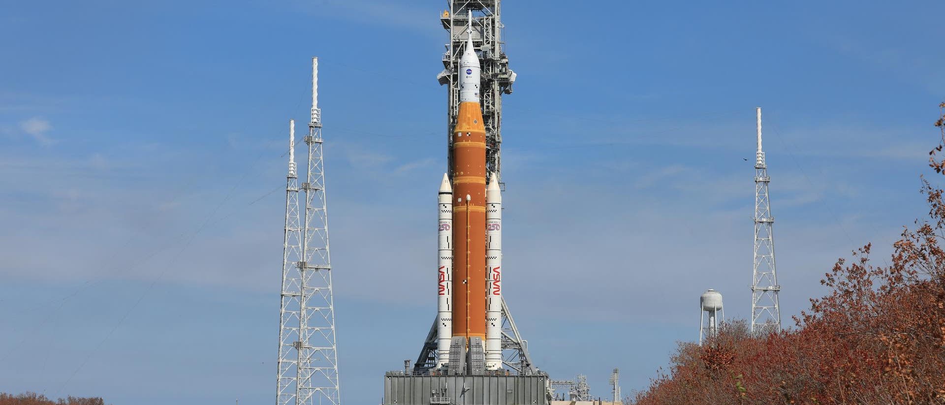Eine gro&szlig;e Rakete steht auf einer Startrampe, umgeben von mehreren hohen Antennenmasten. Der Himmel ist klar und blau. Im Vordergrund ist eine asphaltierte Stra&szlig;e zu sehen, die zur Rampe f&uuml;hrt. Rechts im Bild sind B&uuml;sche mit herbstlich gef&auml;rbtem Laub. Die Rakete ist auf einem mobilen Transporter montiert.