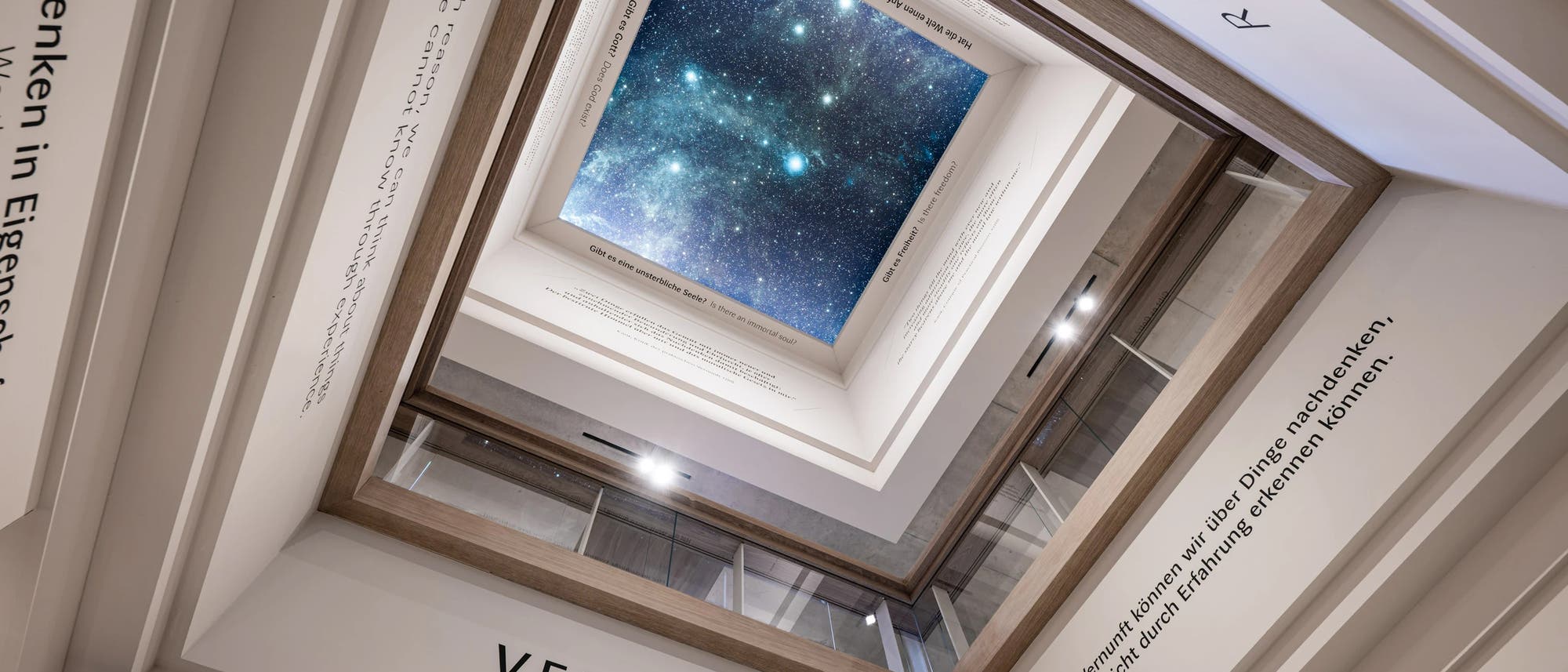 Blick nach oben durch ein mehrst&ouml;ckiges Geb&auml;ude mit einem rechteckigen Lichthof, der einen sternenklaren Nachthimmel zeigt. An den W&auml;nden sind Zitate in verschiedenen Sprachen angebracht, darunter das Wort &bdquo;VERNUNFT&ldquo;. Die Architektur lenkt den Blick auf den Himmel, was eine Verbindung zwischen Wissenschaft und Philosophie suggeriert.