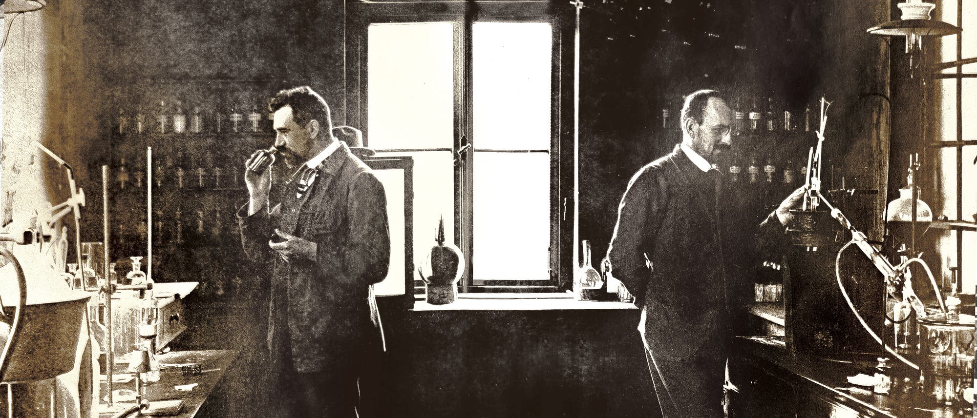 Zwei Männer in einem historischen Labor, umgeben von Glasflaschen und chemischen Apparaturen. Einer der Männer hält ein Objekt in der Hand und scheint es zu untersuchen, während der andere an einem Tisch arbeitet. Im Hintergrund sind Regale mit weiteren Laborutensilien zu sehen. Das Bild vermittelt eine Atmosphäre wissenschaftlicher Forschung aus einer früheren Zeit. Zwei Männer in einem historischen Labor, umgeben von Glasflaschen und chemischen Apparaturen. Einer der Männer hält ein Objekt in der Hand und scheint es zu untersuchen, während der andere an einem Tisch arbeitet. Im Hintergrund sind Regale mit weiteren Laborutensilien zu sehen. Das Bild vermittelt eine Atmosphäre wissenschaftlicher Forschung aus einer früheren Zeit.