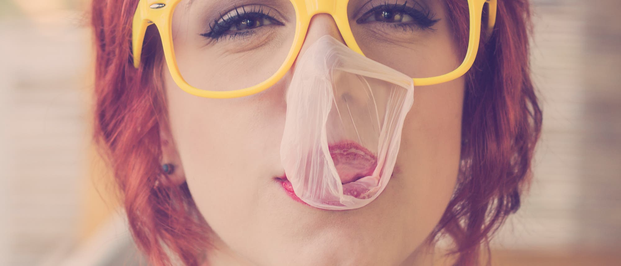 Eine rothaarige junge Frau mit gelber Brille hat eine geplatzte Kuagummiblase im Gesicht.