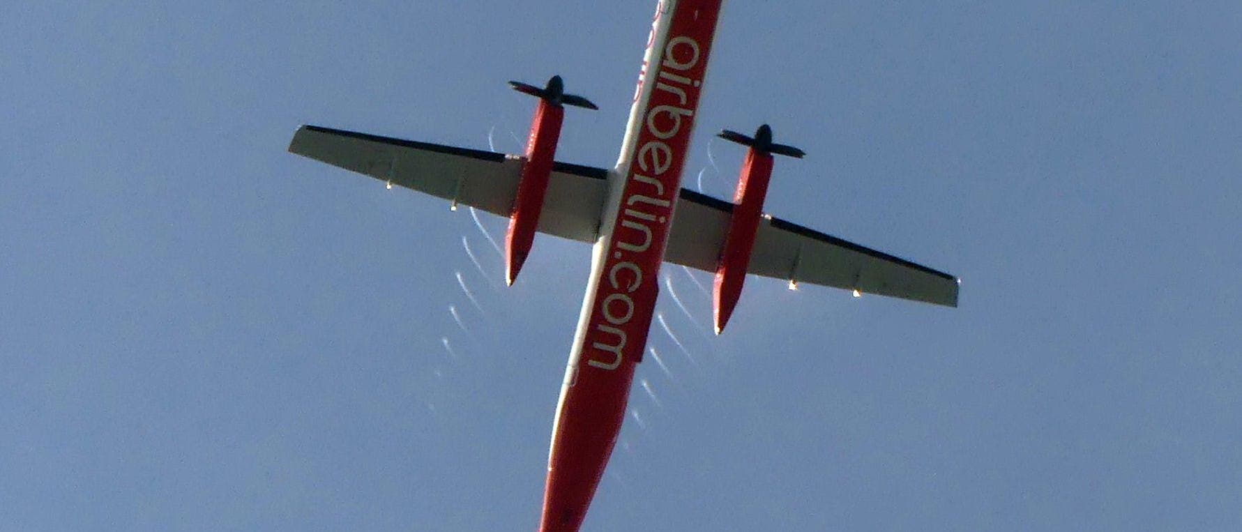 Ein rotes Flugzeug mit der Aufschrift "airberlin.com" auf dem Rumpf fliegt am klaren blauen Himmel. Das Flugzeug ist von unten zu sehen, mit deutlich erkennbaren Tragfl&auml;chen und Propellern. Hinter den Propellern sind kleine, helle Abschnitte von Nebelspiralen zu erkennen, die von Kondenswasser verursacht werden.