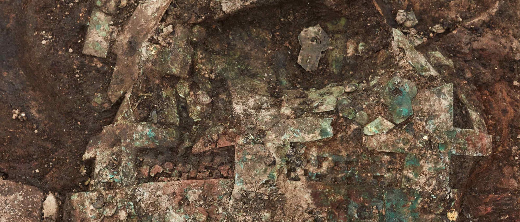 Eine archäologische Ausgrabungsstätte zeigt ein antikes, stark korrodiertes Metallobjekt im Erdreich. Um das Objekt herum sind goldene Schmuckstücke und blaue Perlen verstreut. Eine archäologische Ausgrabungsstätte zeigt ein antikes, stark korrodiertes Metallobjekt im Erdreich. Um das Objekt herum sind goldene Schmuckstücke und blaue Perlen verstreut.