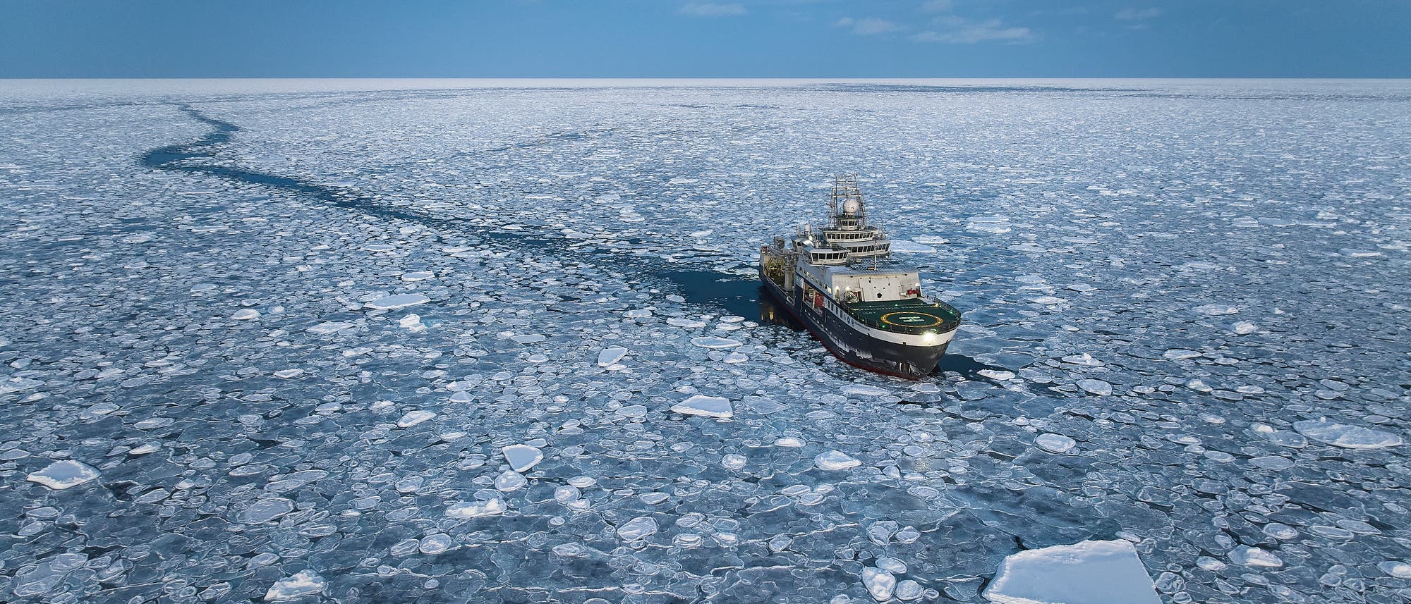 Ein Eisbrecher fährt durch eine weite, mit Eisschollen bedeckte Meeresfläche. Der Himmel ist bewölkt, und der Mond ist sichtbar. Das Schiff hinterlässt eine schmale, eisfreie Spur im Wasser.