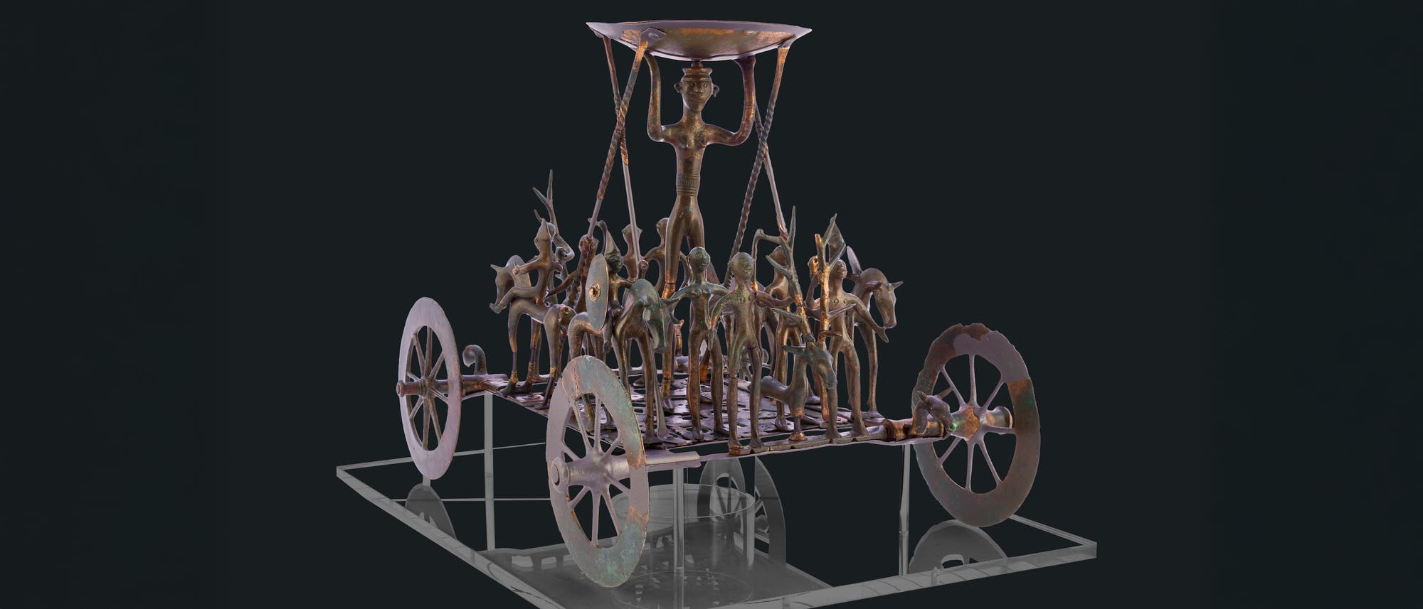 Eine antike bronzene Skulptur, die einen Wagen mit vier großen Rädern darstellt. Auf dem Wagen stehen mehrere kleine Figuren, die Tiere und Menschen darstellen. In der Mitte hält eine größere Figur eine Schale über dem Kopf. Die Skulptur ist detailliert gearbeitet und zeigt kunstvolle Verzierungen. Sie steht auf einer transparenten Plattform, die den Eindruck von Schweben vermittelt. Eine antike bronzene Skulptur, die einen Wagen mit vier großen Rädern darstellt. Auf dem Wagen stehen mehrere kleine Figuren, die Tiere und Menschen darstellen. In der Mitte hält eine größere Figur eine Schale über dem Kopf. Die Skulptur ist detailliert gearbeitet und zeigt kunstvolle Verzierungen. Sie steht auf einer transparenten Plattform, die den Eindruck von Schweben vermittelt.