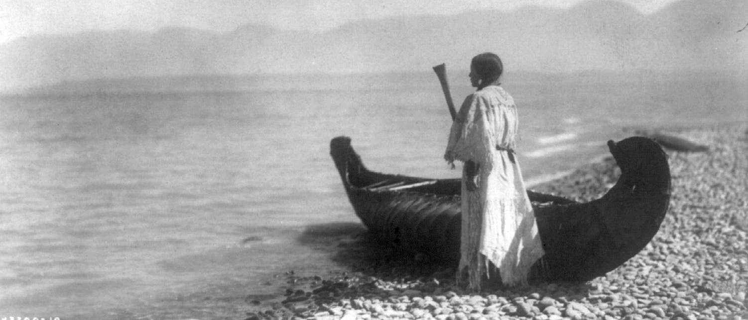 Eine Angehörige der Kutenai steht um 1910 neben einem Kanu in Nordamerika.