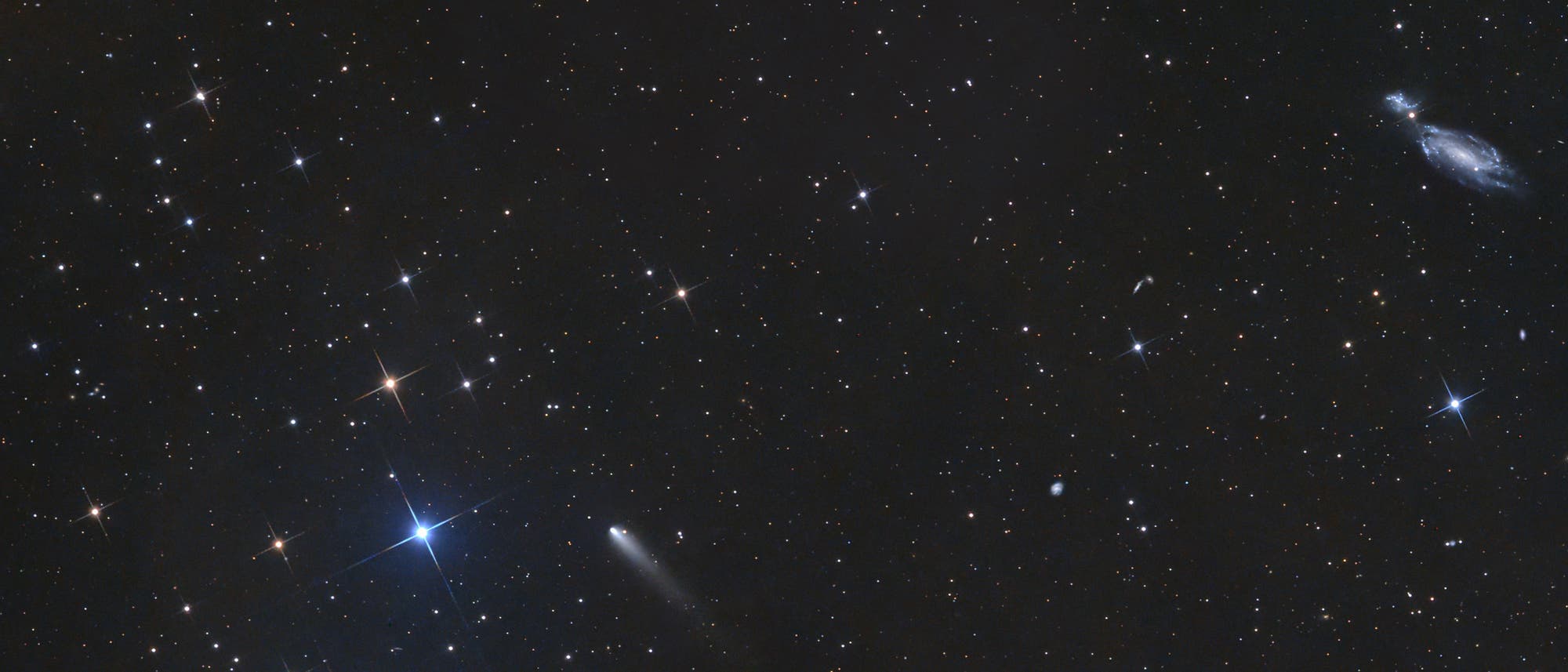 Ein astronomisches Bild zeigt einen sternen&uuml;bers&auml;ten Nachthimmel. Mehrere helle Sterne sind sichtbar, einige mit deutlichen Lichtstrahlen. In der unteren Bildmitte ist ein Komet mit einem leuchtenden Schweif zu sehen. Rechts oben befindet sich eine Spiralgalaxie mit klar erkennbaren Armen. Der Hintergrund ist dunkel, was die Helligkeit der Sterne und des Kometen betont.