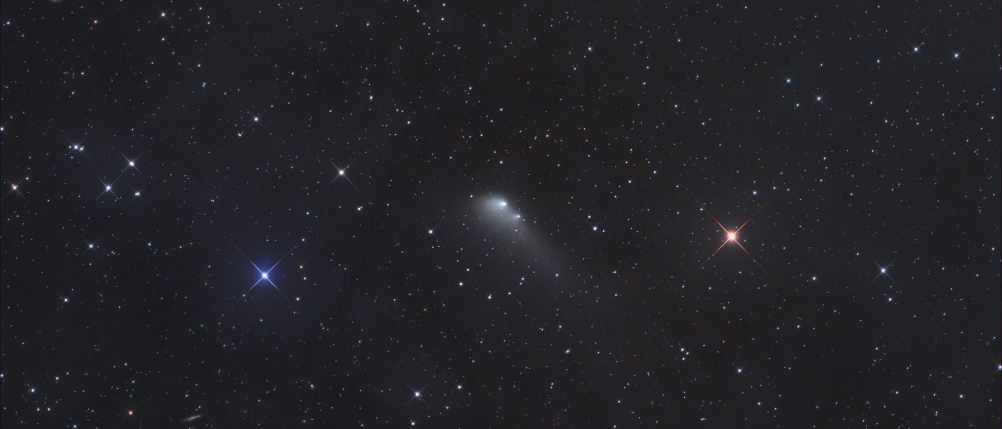 Ein astronomisches Bild zeigt einen Nachthimmel voller Sterne. In der Mitte ist ein Komet mit einem leuchtenden Kern und einem diffusen Schweif zu sehen, der nach rechts unten zeigt. Der Hintergrund ist mit zahlreichen funkelnden Sternen unterschiedlicher Helligkeit übersät. Einige Sterne erscheinen in verschiedenen Farben, darunter ein auffälliger blauer Stern links und ein rötlicher Stern rechts. Der Himmel ist dunkel und klar, was die Details des Kometen und der Sterne hervorhebt. Ein astronomisches Bild zeigt einen Nachthimmel voller Sterne. In der Mitte ist ein Komet mit einem leuchtenden Kern und einem diffusen Schweif zu sehen, der nach rechts unten zeigt. Der Hintergrund ist mit zahlreichen funkelnden Sternen unterschiedlicher Helligkeit übersät. Einige Sterne erscheinen in verschiedenen Farben, darunter ein auffälliger blauer Stern links und ein rötlicher Stern rechts. Der Himmel ist dunkel und klar, was die Details des Kometen und der Sterne hervorhebt.
