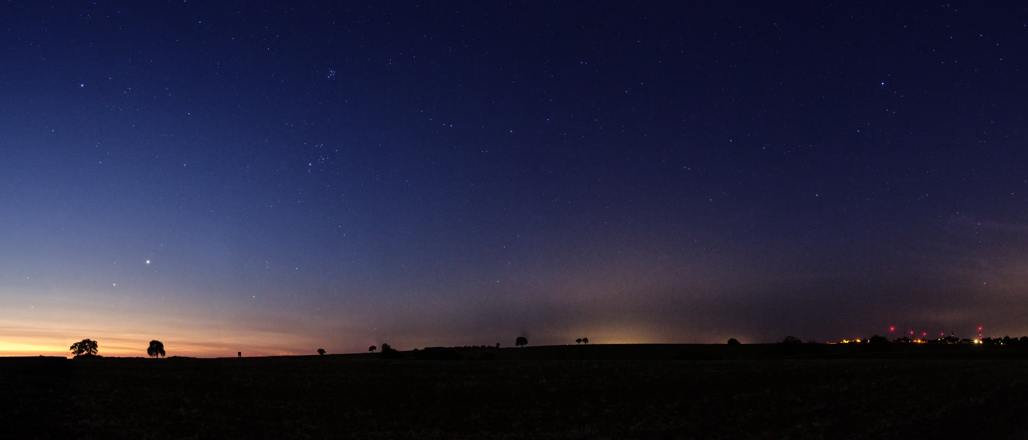 Eine nächtliche Landschaft mit einem klaren Sternenhimmel. Am Horizont sind Silhouetten von Bäumen und eine schwache Lichtverschmutzung von einer Stadt zu sehen. Der Himmel zeigt zahlreiche Sterne, die in der Dunkelheit leuchten. Links ist ein schwacher Sonnenaufgang zu erkennen, der den Himmel in warmen Farben färbt. Eine nächtliche Landschaft mit einem klaren Sternenhimmel. Am Horizont sind Silhouetten von Bäumen und eine schwache Lichtverschmutzung von einer Stadt zu sehen. Der Himmel zeigt zahlreiche Sterne, die in der Dunkelheit leuchten. Links ist ein schwacher Sonnenaufgang zu erkennen, der den Himmel in warmen Farben färbt.