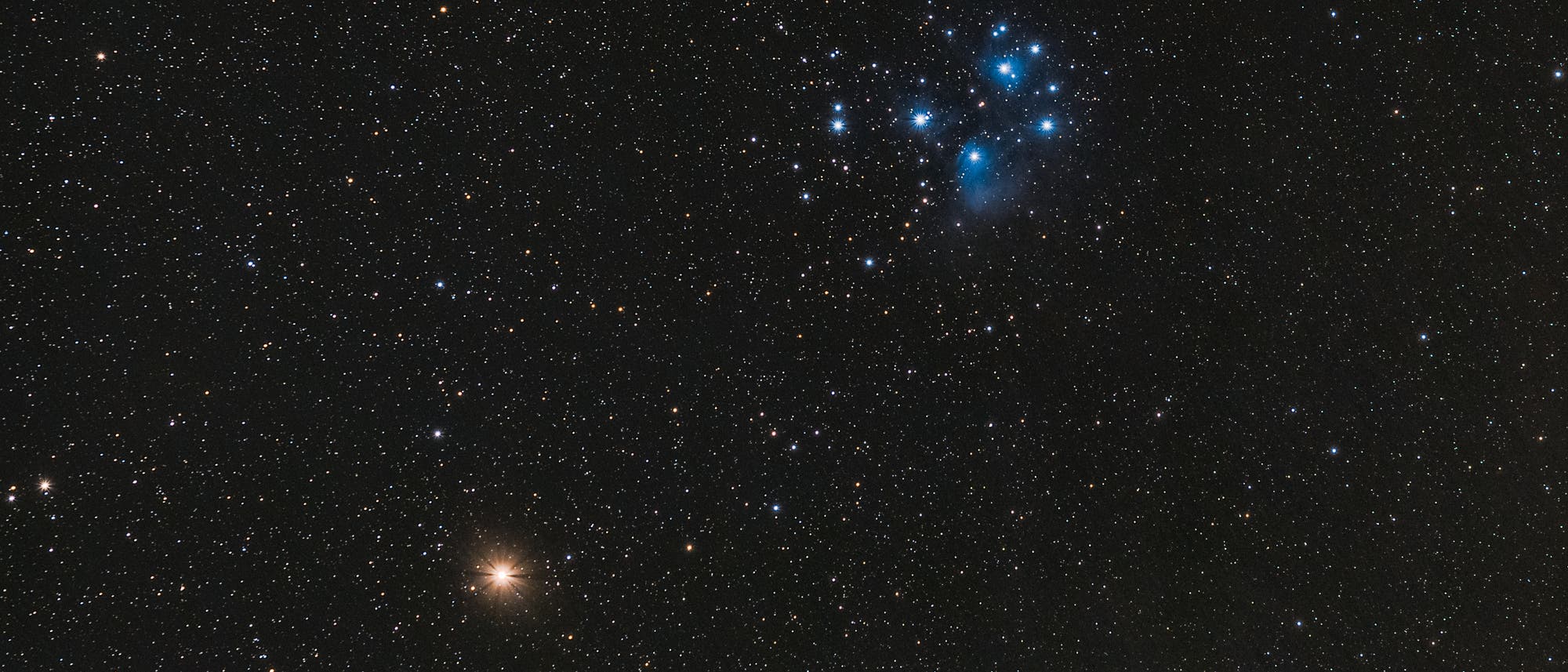 Ein Sternenhimmel mit zahlreichen leuchtenden Punkten vor einem dunklen Hintergrund. Rechts oben ist eine Ansammlung von bl&auml;ulich leuchtenden Sternen zu sehen, die als Sternhaufen erscheinen. Links unten befindet sich ein heller, orangefarbener Stern. Das Bild zeigt die Weite des Universums und die Vielfalt der Sterne.