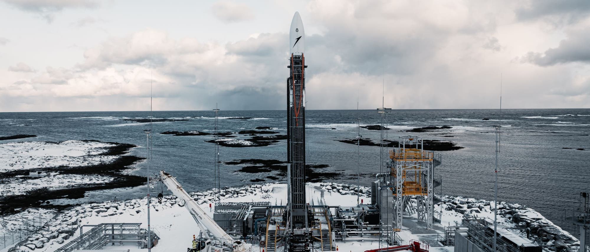 Eine Rakete steht auf einer verschneiten Startrampe an der Küste, umgeben von technischen Anlagen und Containern. Im Hintergrund ist das Meer mit kleinen Inseln zu sehen, darüber ein bewölkter Himmel. Die Szene vermittelt eine ruhige, aber technisch fortschrittliche Atmosphäre. Eine Rakete steht auf einer verschneiten Startrampe an der Küste, umgeben von technischen Anlagen und Containern. Im Hintergrund ist das Meer mit kleinen Inseln zu sehen, darüber ein bewölkter Himmel. Die Szene vermittelt eine ruhige, aber technisch fortschrittliche Atmosphäre.