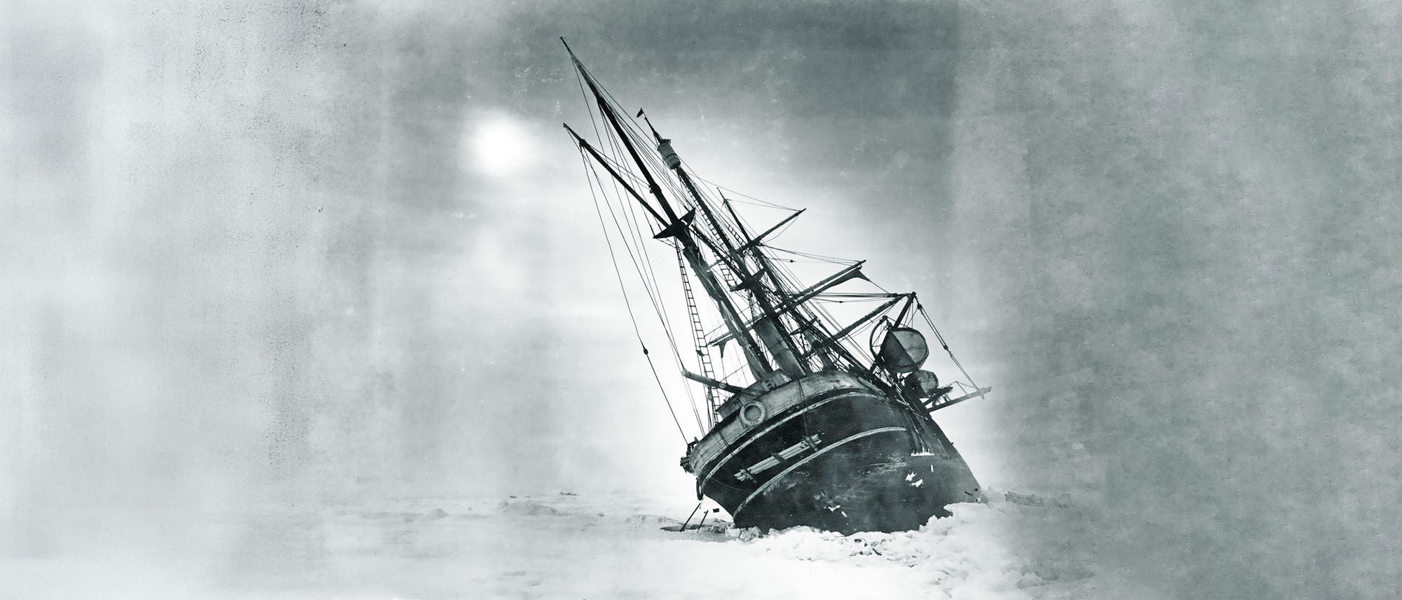Ein historisches Schwarz-Wei&szlig;-Foto zeigt ein gro&szlig;es Segelschiff, das im Eis gefangen ist. Das Schiff ist zur Seite geneigt, umgeben von einer weiten, eisigen Landschaft. Der Himmel ist bew&ouml;lkt, was die dramatische und herausfordernde Situation des Schiffes unterstreicht. Die Masten und Takelage sind deutlich sichtbar, was auf die Gr&ouml;&szlig;e und Komplexit&auml;t des Schiffes hinweist.