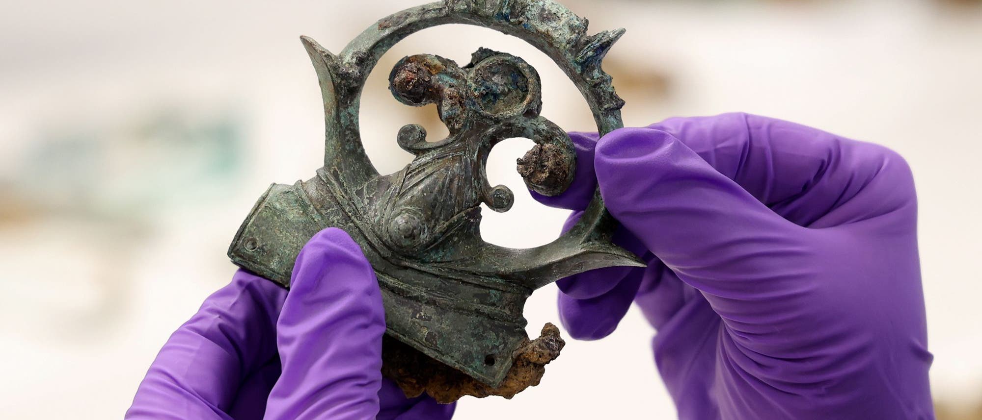 Eine Person mit lila Handschuhen hält ein antikes, verziertes Metallartefakt, das möglicherweise Teil eines historischen Fundes ist. Das Artefakt hat eine komplexe, dekorative Struktur mit oxidierten und korrodierten Stellen. Der Hintergrund ist unscharf, was den Fokus auf das Artefakt lenkt. Eine Person mit lila Handschuhen hält ein antikes, verziertes Metallartefakt, das möglicherweise Teil eines historischen Fundes ist. Das Artefakt hat eine komplexe, dekorative Struktur mit oxidierten und korrodierten Stellen. Der Hintergrund ist unscharf, was den Fokus auf das Artefakt lenkt.