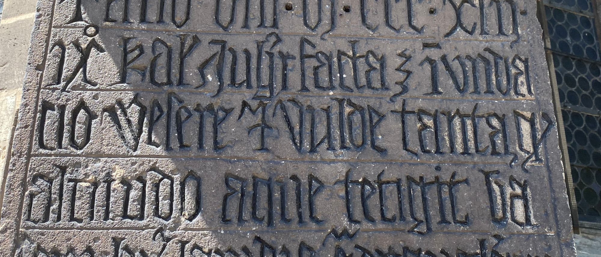 Eine Steinplatte mit eingraviertem Text in gotischer Schrift. Der Text ist in mehreren Zeilen angeordnet und enthält historische oder religiöse Inschriften. Die Buchstaben sind kunstvoll gestaltet und in den Stein eingraviert, was auf eine alte oder mittelalterliche Herkunft hindeutet. Die Platte ist Teil einer größeren architektonischen Struktur. Eine Steinplatte mit eingraviertem Text in gotischer Schrift. Der Text ist in mehreren Zeilen angeordnet und enthält historische oder religiöse Inschriften. Die Buchstaben sind kunstvoll gestaltet und in den Stein eingraviert, was auf eine alte oder mittelalterliche Herkunft hindeutet. Die Platte ist Teil einer größeren architektonischen Struktur.