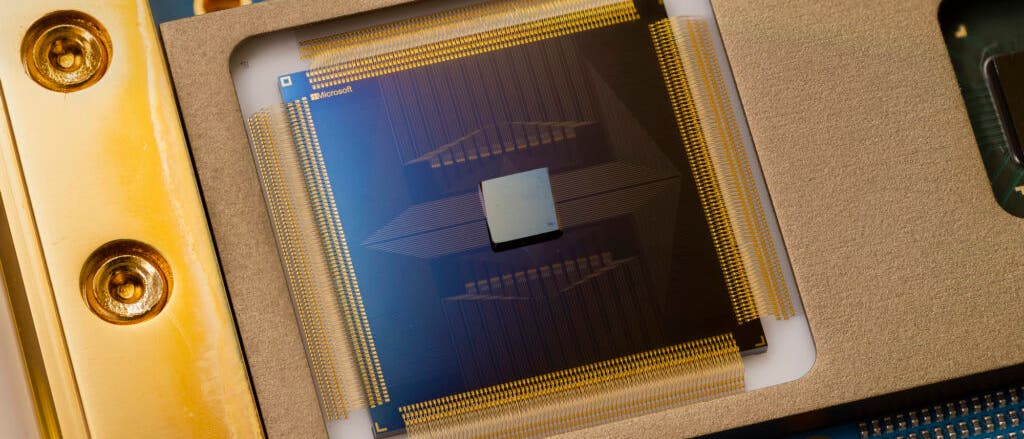 Nahaufnahme eines Computerchips, eingebettet in eine goldene Halterung. Der Chip zeigt feine, goldene Leiterbahnen und ein zentrales quadratisches Element. Im Hintergrund sind Teile einer blauen Platine mit goldenen Schrauben sichtbar. Oben links ist ein kleines Logo mit dem Text "Microsoft" zu erkennen.