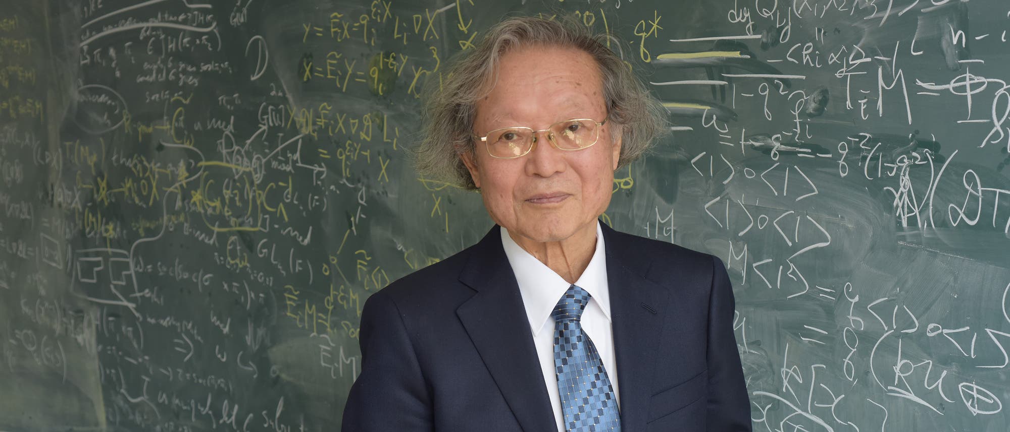 Der Abelpreisträger Masaki Kashiwara steht in einem dunkelblauen Anzug mit Krawatte vor einer Tafel voller mathematischen Formeln. Er hält ein gelbes Buch mit dem Titel »Nobel Lectures in Physics 1971-1980« in den Händen und schaut direkt in die Kamera. Der Abelpreisträger Masaki Kashiwara steht in einem dunkelblauen Anzug mit Krawatte vor einer Tafel voller mathematischen Formeln. Er hält ein gelbes Buch mit dem Titel »Nobel Lectures in Physics 1971-1980« in den Händen und schaut direkt in die Kamera.