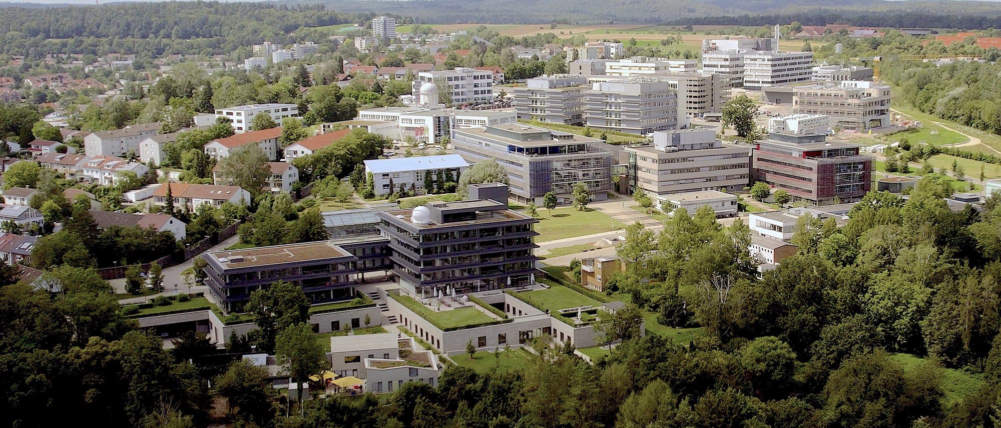Luftaufnahme eines Universit&auml;tscampus, umgeben von gr&uuml;nen W&auml;ldern und einer kleinen Stadt. Mehrere moderne Geb&auml;ude mit Flachd&auml;chern und Glasfassaden sind sichtbar, eingebettet in eine Landschaft aus B&auml;umen und Rasenfl&auml;chen. Im Hintergrund erstrecken sich H&uuml;gel und Felder unter einem leicht bew&ouml;lkten Himmel.