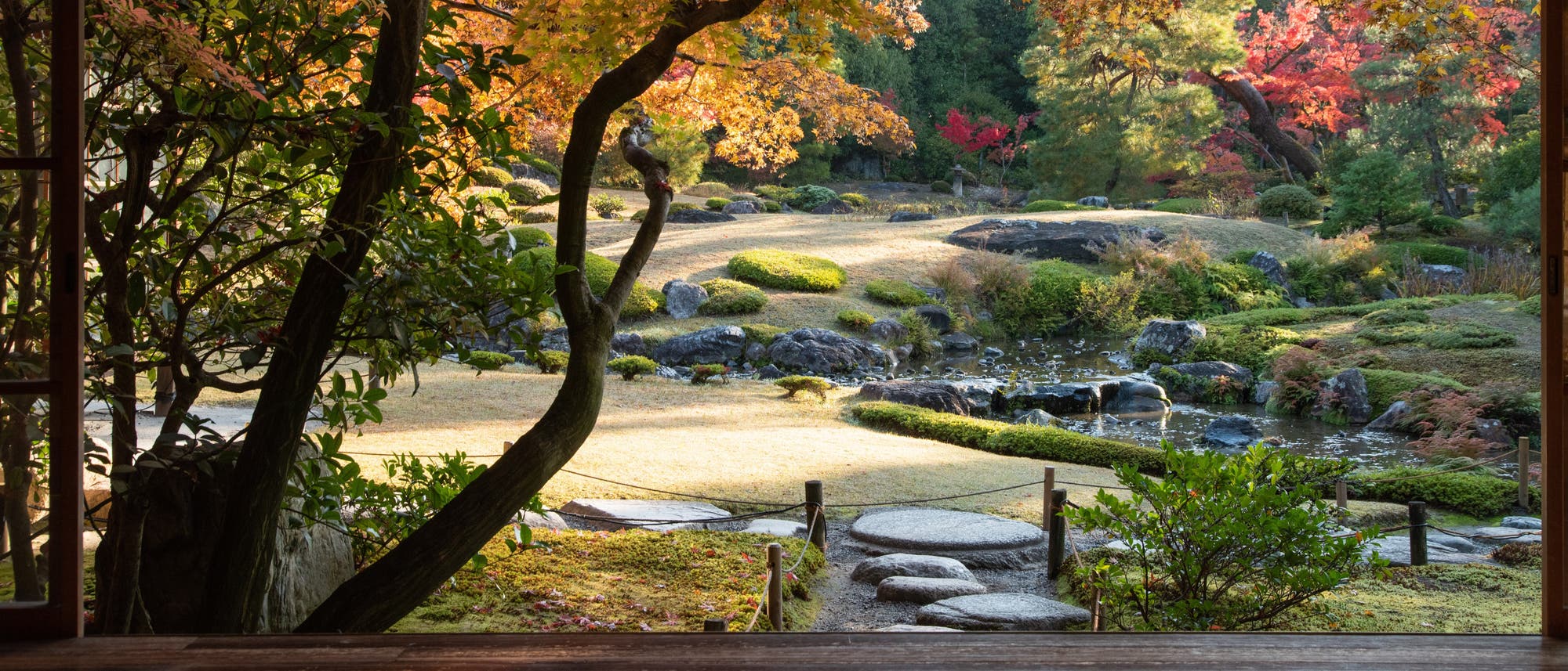 Ein traditioneller japanischer Garten, gesehen durch einen offenen Holzrahmen. Im Vordergrund steht ein Baum mit herbstlich gefärbten Blättern in Orange- und Gelbtönen. Der Garten zeigt gepflegte Rasenflächen, Trittsteine und einen kleinen Bach, umgeben von üppigem Grün und roten Ahornbäumen. Die Szene strahlt Ruhe und natürliche Schönheit aus. Ein traditioneller japanischer Garten, gesehen durch einen offenen Holzrahmen. Im Vordergrund steht ein Baum mit herbstlich gefärbten Blättern in Orange- und Gelbtönen. Der Garten zeigt gepflegte Rasenflächen, Trittsteine und einen kleinen Bach, umgeben von üppigem Grün und roten Ahornbäumen. Die Szene strahlt Ruhe und natürliche Schönheit aus.