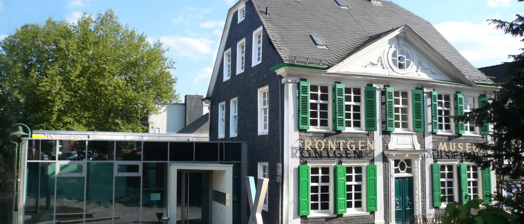 Historisches Gebäude des Röntgen-Museums in Remscheid-Lennep, Deutschland. Das Fachwerkhaus hat grüne Fensterläden und ein graues Schieferdach. Links ist ein moderner Anbau mit Glasfassade zu sehen. Vor dem Eingang steht eine Skulptur in Form eines großen "X". Das Museum ist dem Physiker Wilhelm Conrad Röntgen gewidmet. Historisches Gebäude des Röntgen-Museums in Remscheid-Lennep, Deutschland. Das Fachwerkhaus hat grüne Fensterläden und ein graues Schieferdach. Links ist ein moderner Anbau mit Glasfassade zu sehen. Vor dem Eingang steht eine Skulptur in Form eines großen "X". Das Museum ist dem Physiker Wilhelm Conrad Röntgen gewidmet.