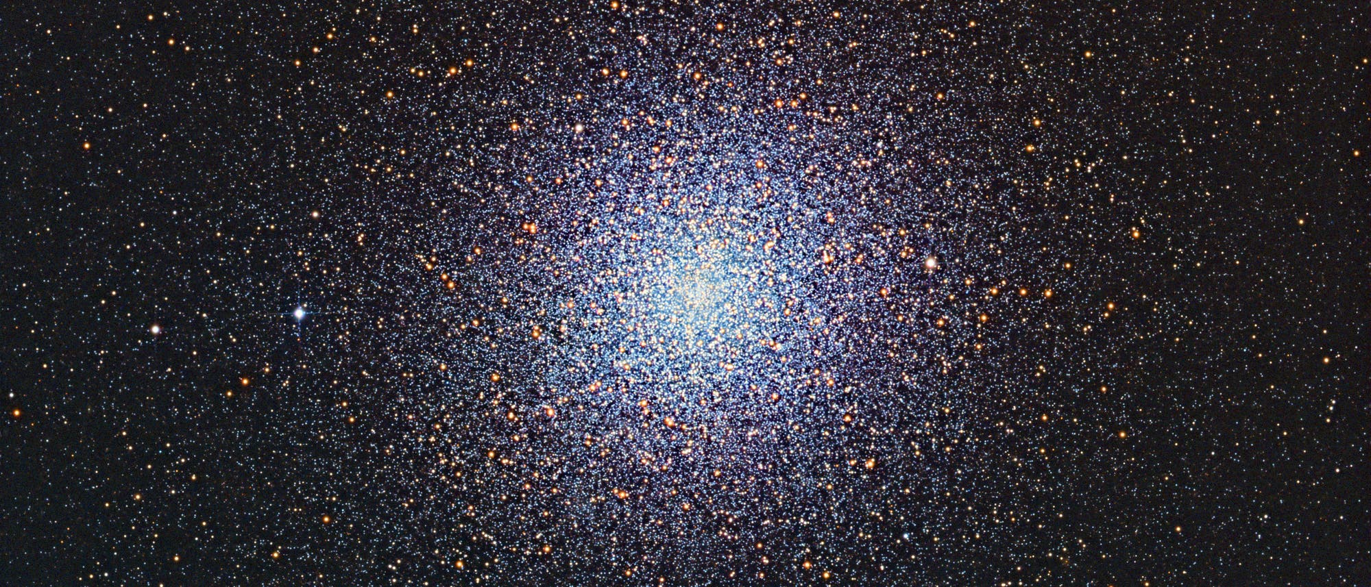 Ein dichtes, kugelf&ouml;rmiges Sternenfeld im Weltraum, das einen Kugelsternhaufen darstellt. Die Sterne sind in verschiedenen Farben sichtbar, haupts&auml;chlich in Blau- und Rott&ouml;nen, und konzentrieren sich in der Mitte des Bildes, was auf eine hohe Sterndichte hinweist. Der Hintergrund ist dunkel, was den Kontrast zu den leuchtenden Sternen verst&auml;rkt.