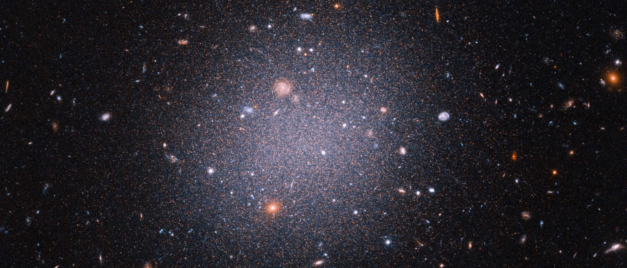 Eine Aufnahme des Weltraums, die eine Vielzahl von Galaxien und Sternen zeigt. Im Zentrum befindet sich eine Ansammlung von Sternen, die von zahlreichen kleineren Galaxien umgeben ist. Die Farben variieren von bl&auml;ulich wei&szlig; bis r&ouml;tlich, was auf unterschiedliche Temperaturen und Entfernungen hinweist. Der Hintergrund ist dunkel, was den Kontrast zu den leuchtenden Himmelsk&ouml;rpern verst&auml;rkt.