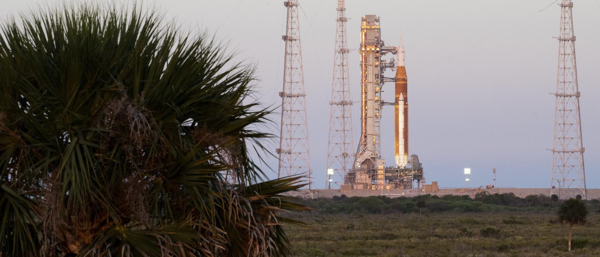 Eine Rakete steht auf einer Startrampe, umgeben von vier hohen Antennenstrukturen. Im Vordergrund ist eine Palme zu sehen. Der Himmel ist klar, und die Szene ist in sanftes Abendlicht getaucht. Die Umgebung ist flach und bewachsen.
