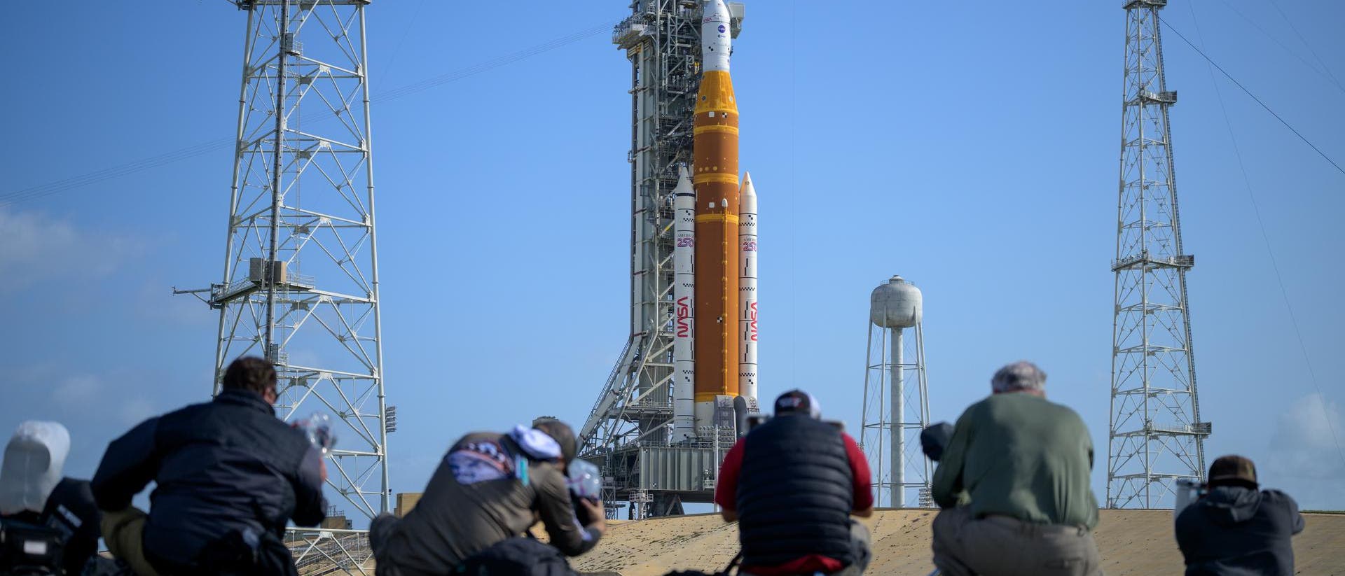 Eine Gruppe von Fotografen kniet auf dem Boden und richtet ihre Kameras auf eine gro&szlig;e Rakete, die auf einer Startrampe steht. Die Rakete ist orange-wei&szlig; und tr&auml;gt das NASA-Logo. Im Hintergrund sind der blaue Himmel und mehrere hohe Metallstrukturen zu sehen, die die Startrampe umgeben.